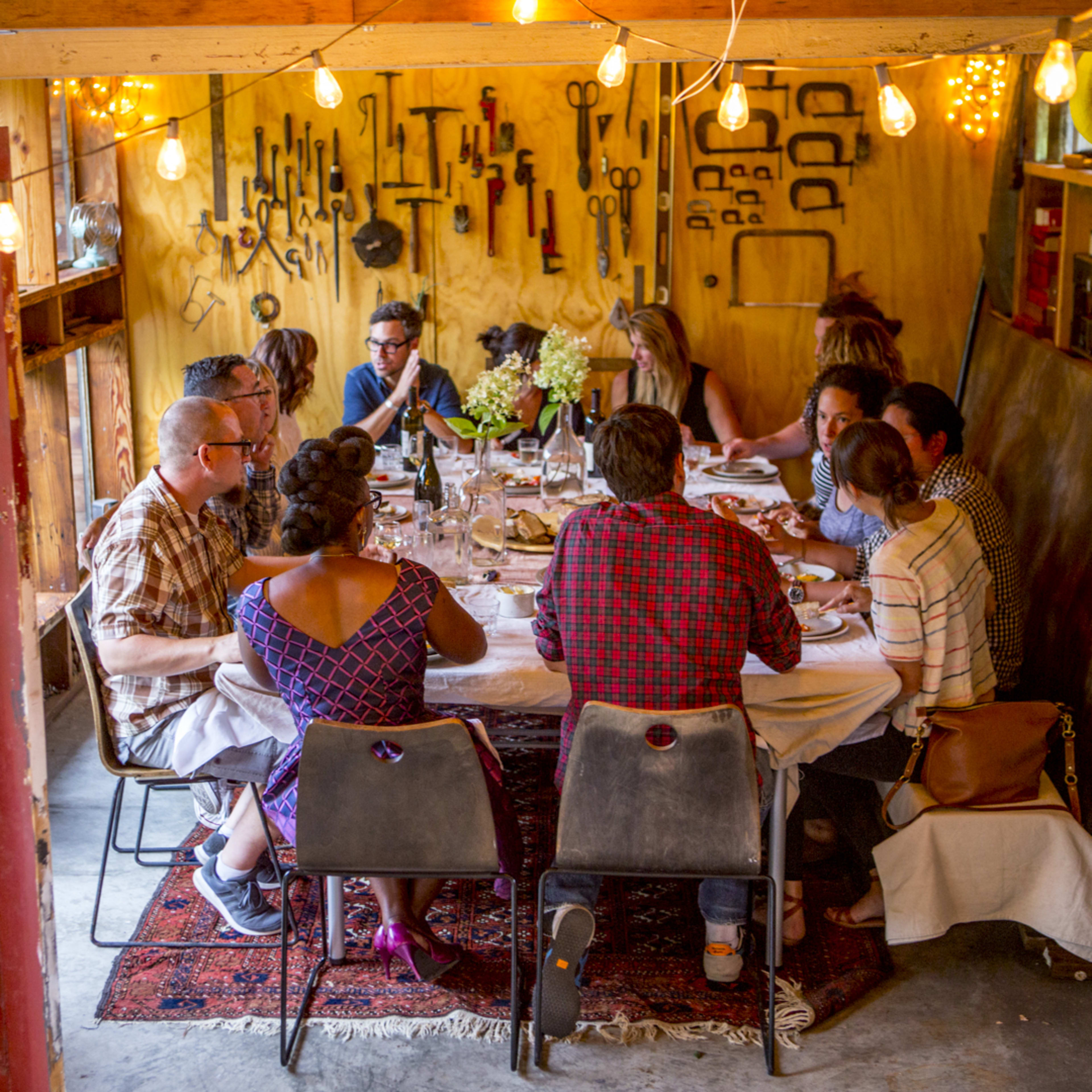A group of people sits around a large table in a rustic room decorated with various tools and pendant lights.