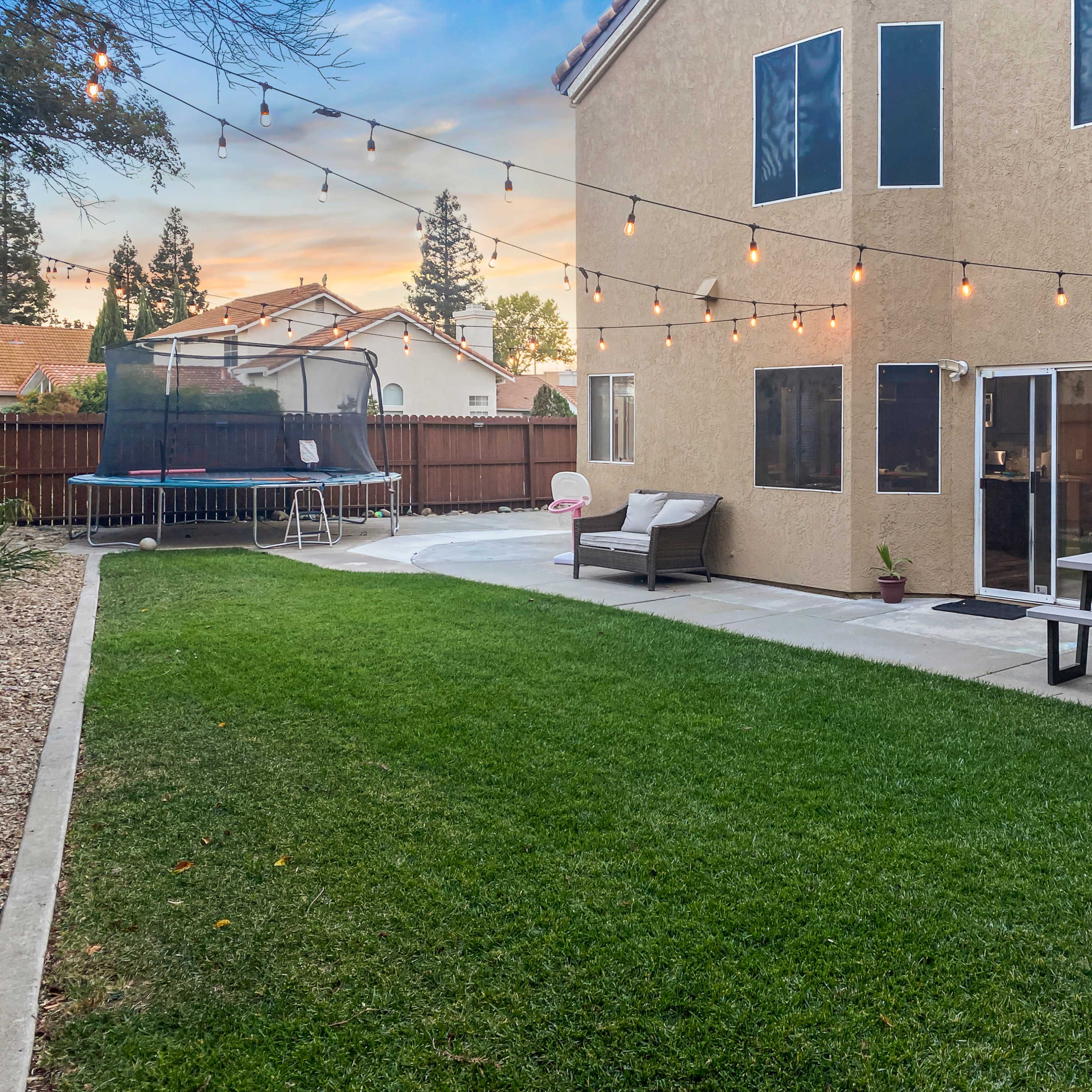 A backyard with a green lawn, a trampoline, a patio set, and string lights hanging overhead, leading to the house on one side.