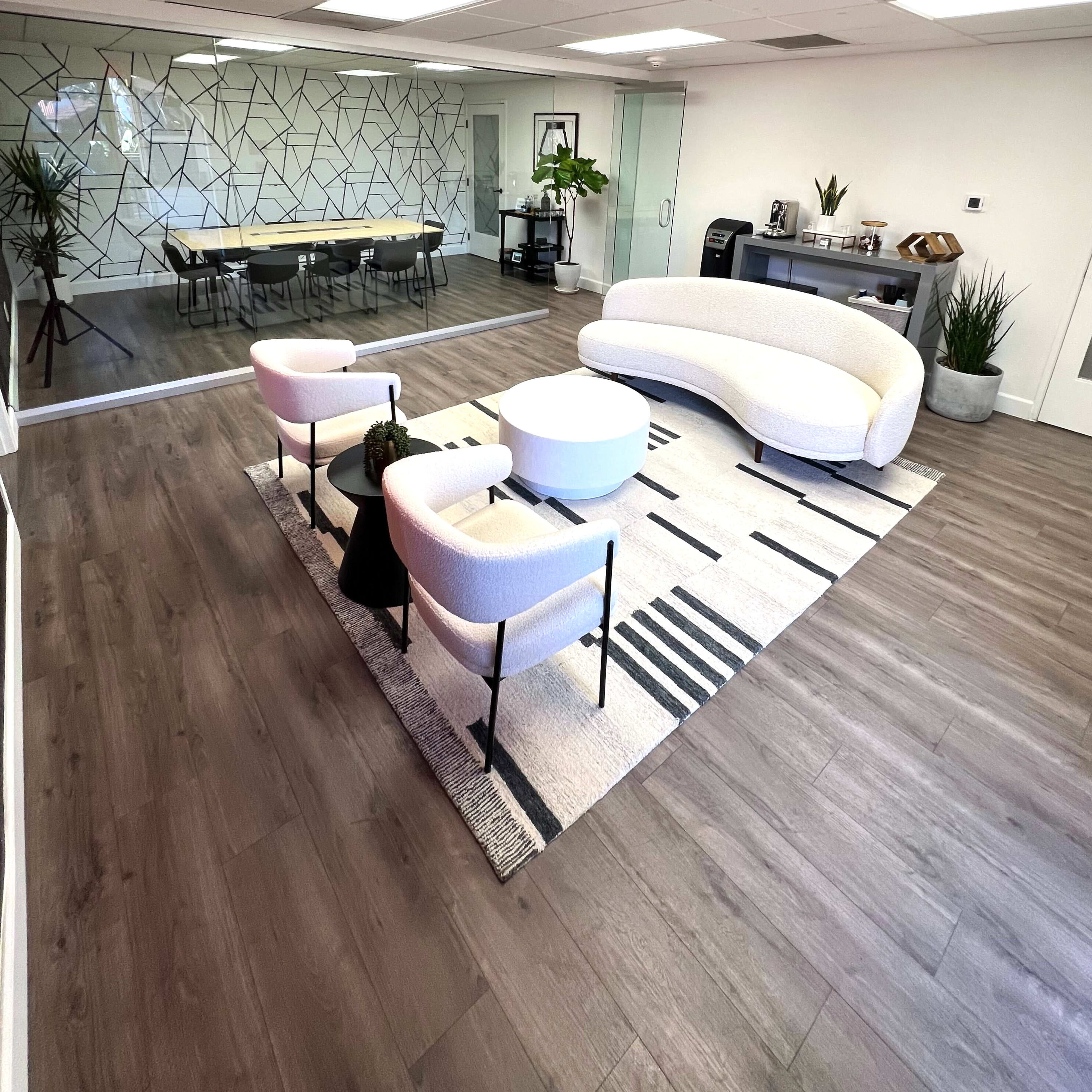 A modern office space features a curved white sofa, two accent chairs, and a round coffee table on a patterned rug, with a conference table in the background and greenery throughout.