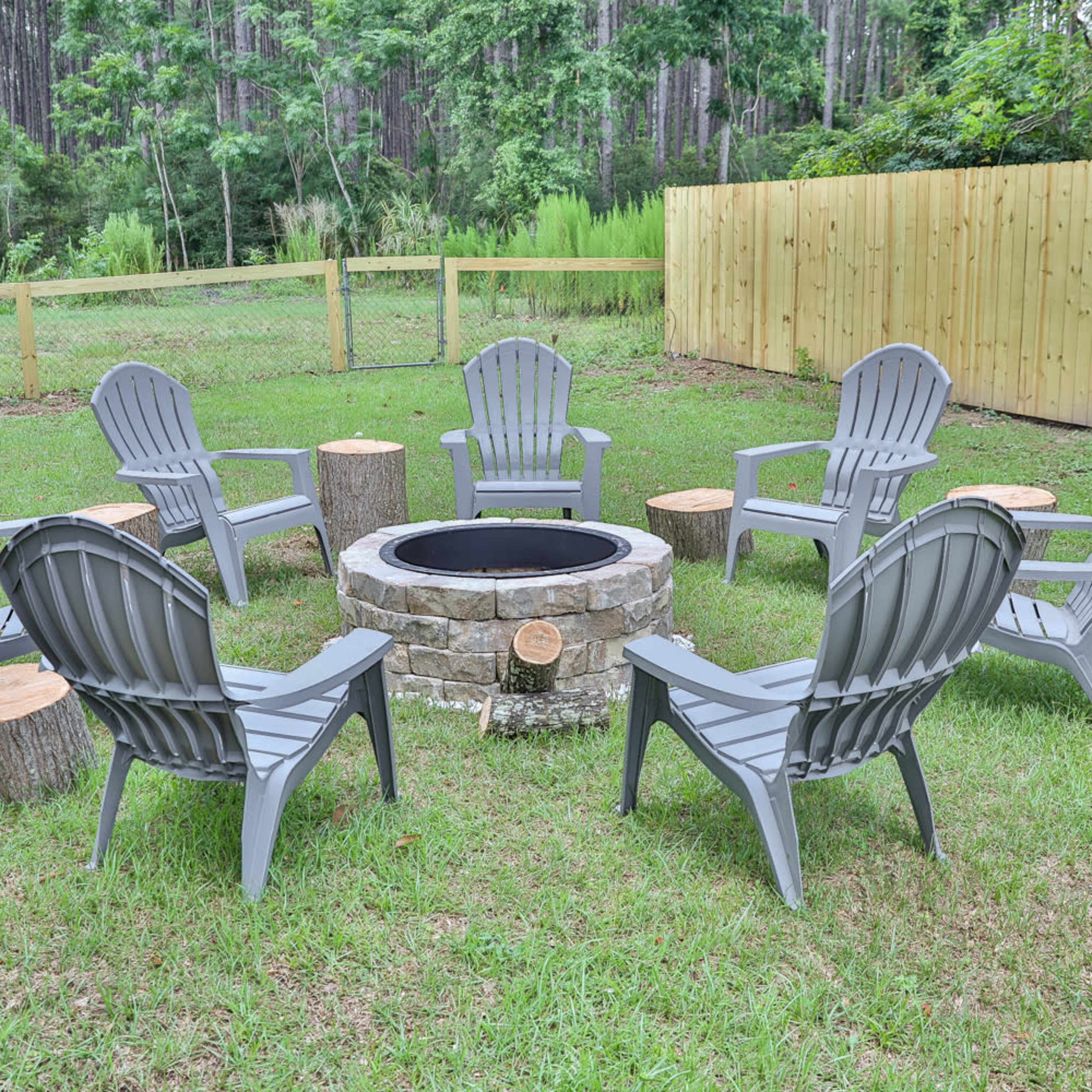 A circular arrangement of six gray Adirondack chairs surrounds a stone fire pit in a grassy area, bordered by a wooden fence and tall trees.