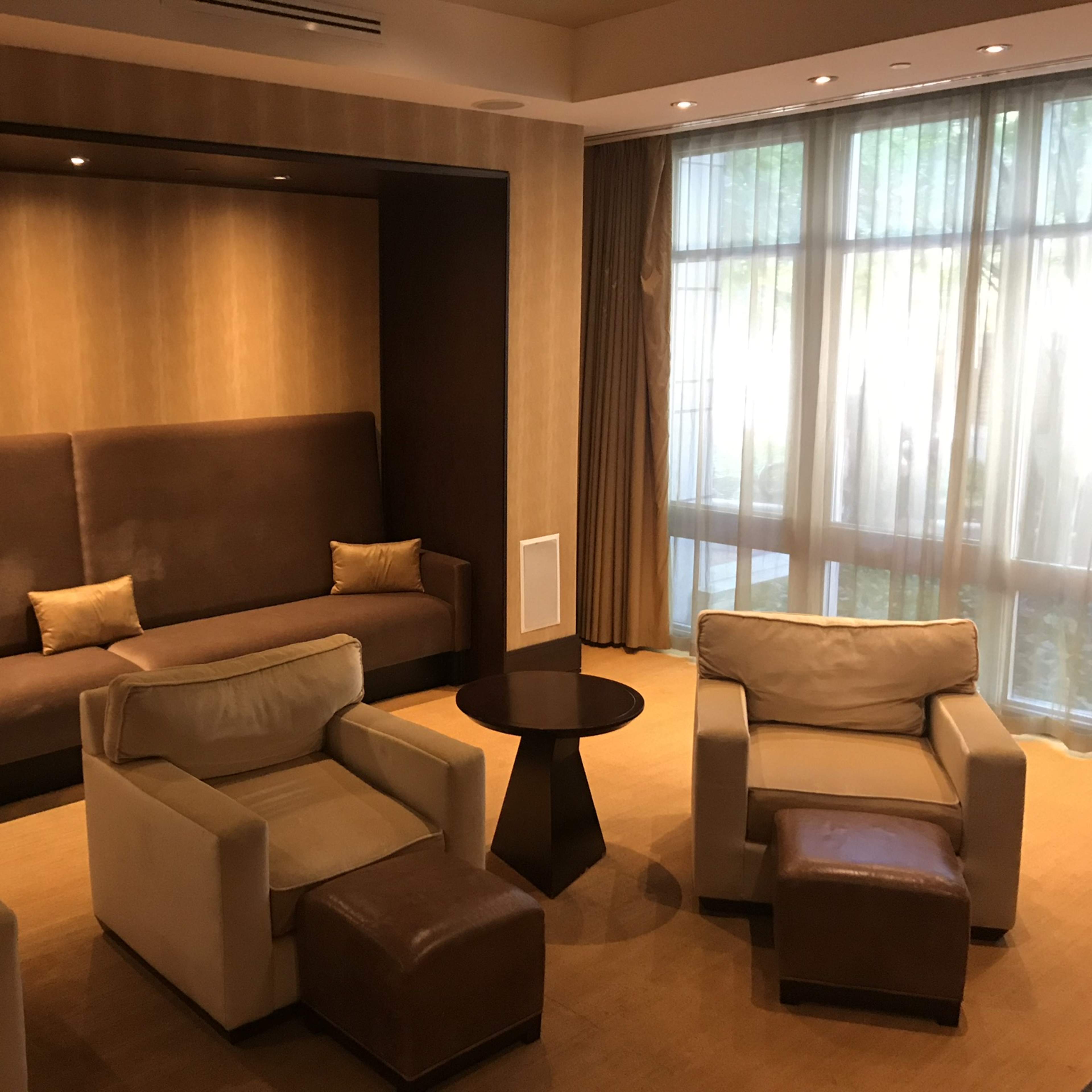 A lounge area with a brown upholstered wall, two light-colored armchairs, a small round table, and a beige sofa positioned against a backdrop of large windows covered by sheer curtains.