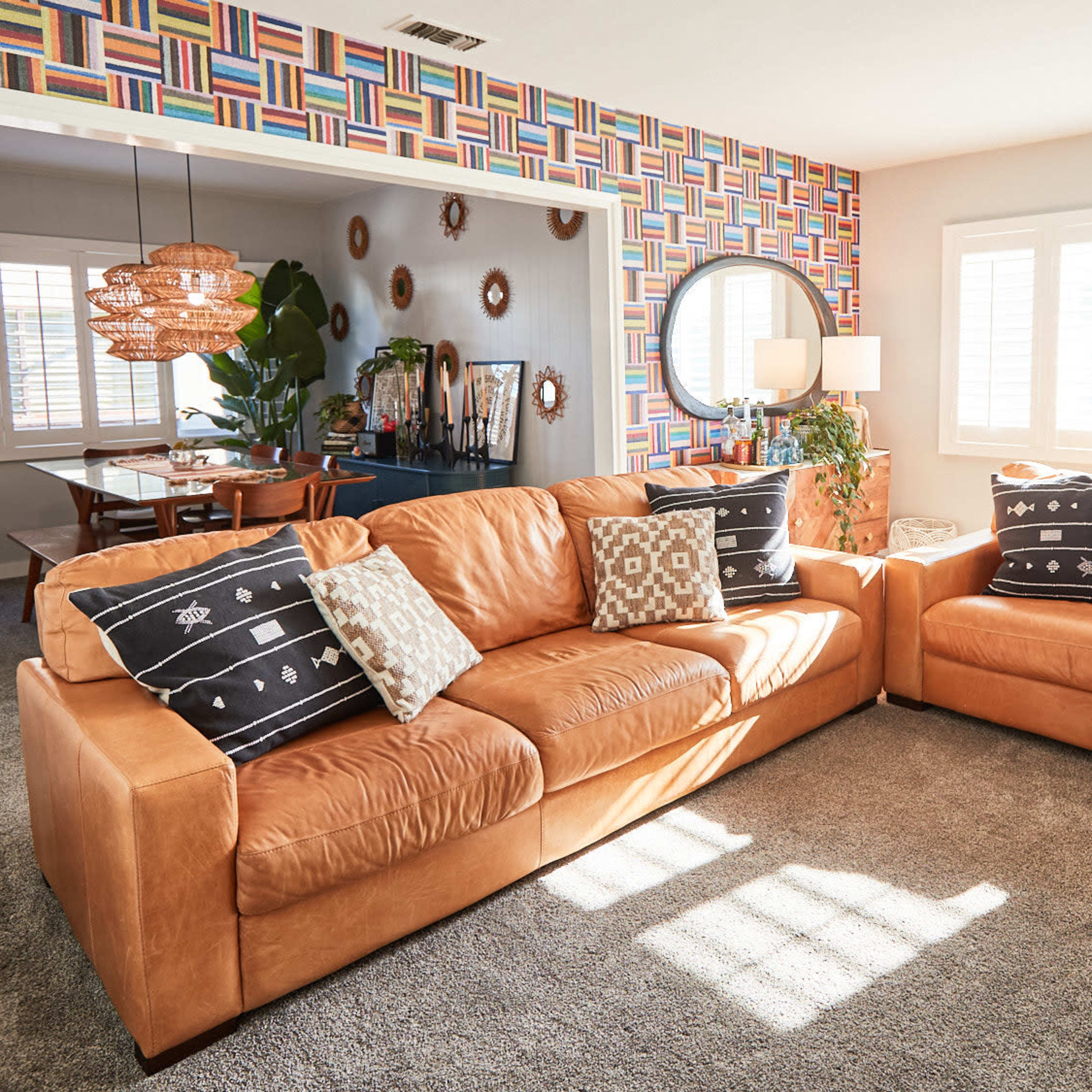 A living room with tan leather sofas, a round mirror on the wall, and colorful patterned wallpaper, illuminated by natural light from the windows.