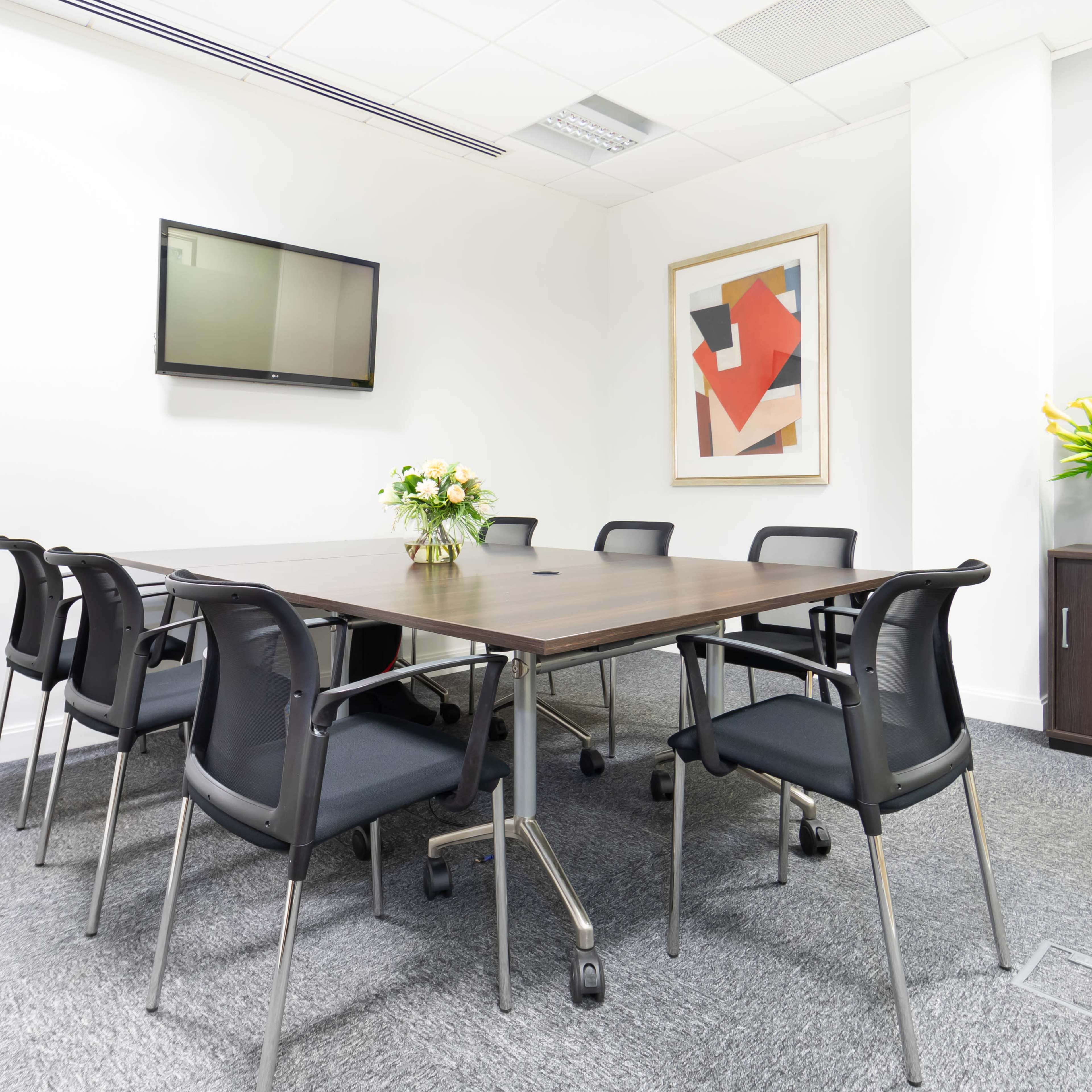 A conference room features a large rectangular table surrounded by six black chairs, a wall-mounted television, and a floral arrangement on the table.