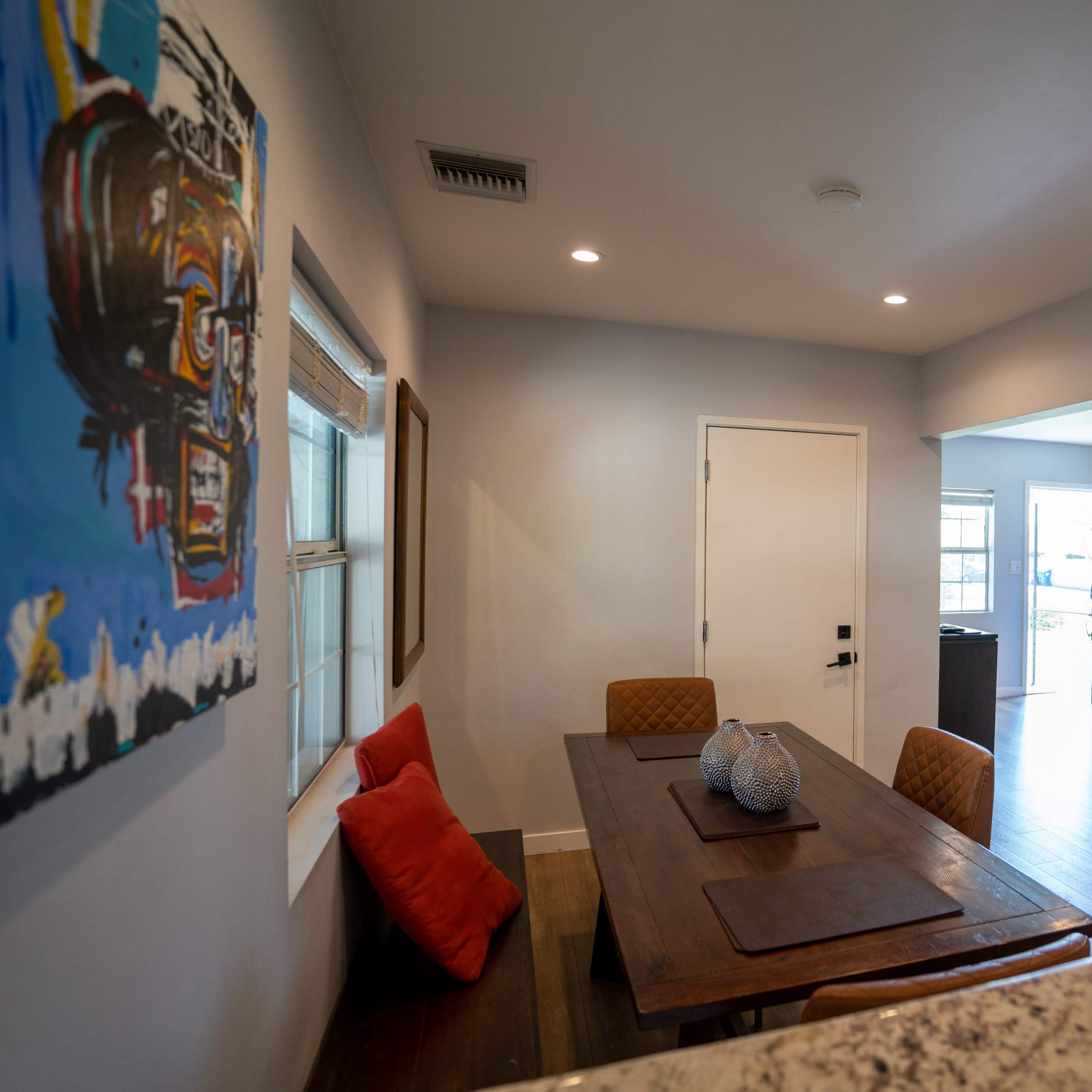 A modern interior space featuring a dining table with chairs, colorful artwork on the wall, and a view of a living area beyond.