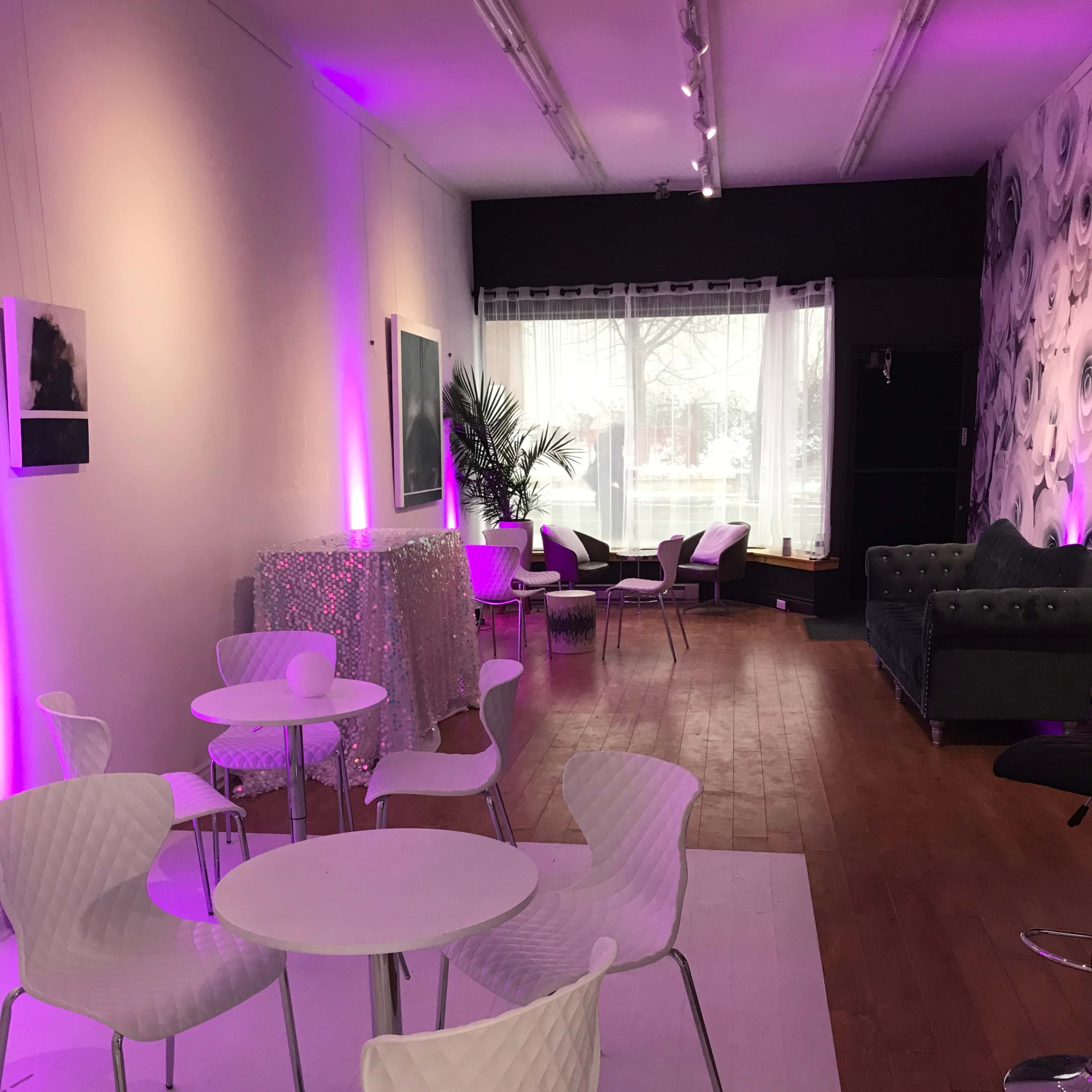 A stylish interior space with white tables and chairs, purple lighting, and a mix of seating areas, including a bar and a plush couch.