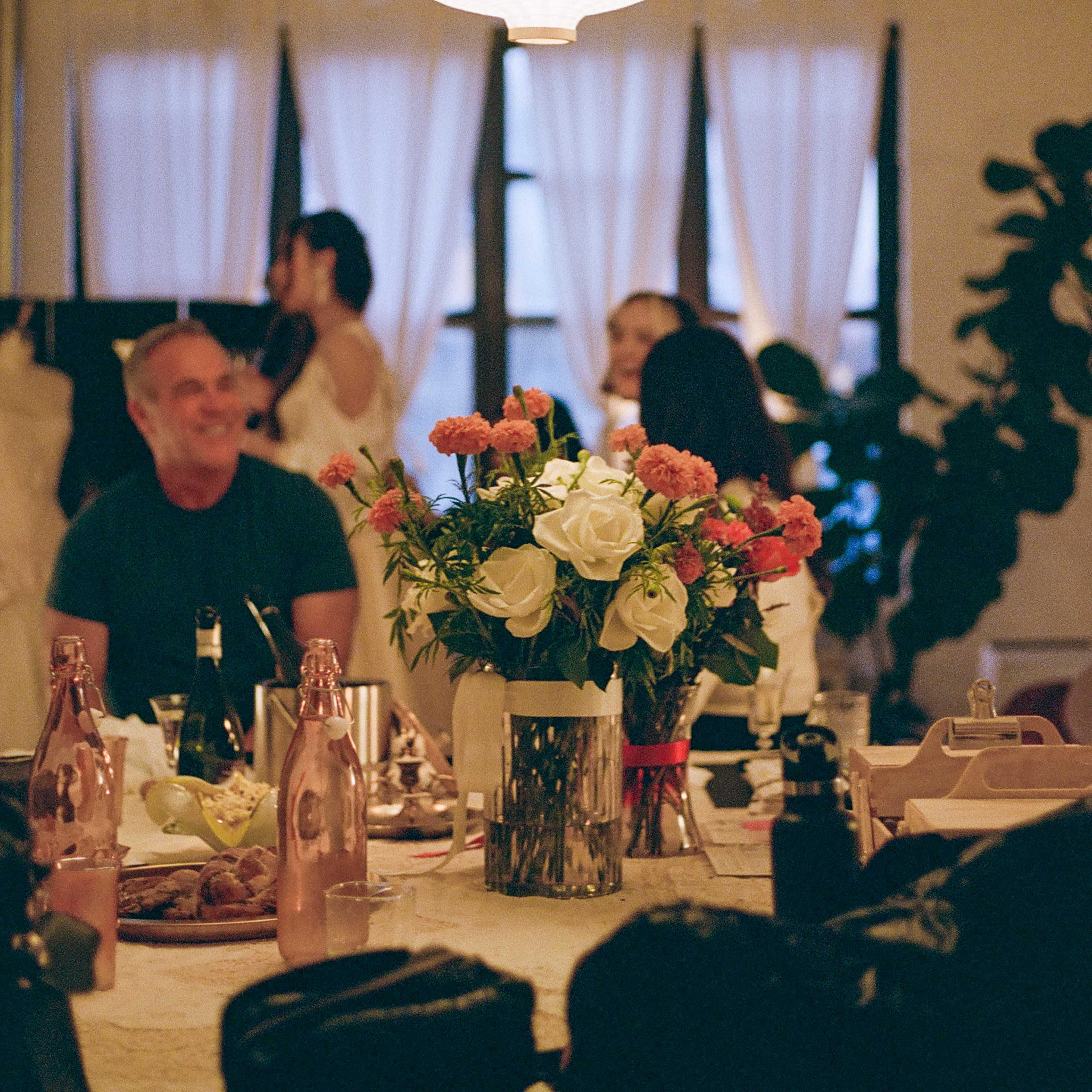 A round paper lantern hangs above a table set with flowers, while people are gathered around in a softly lit room.