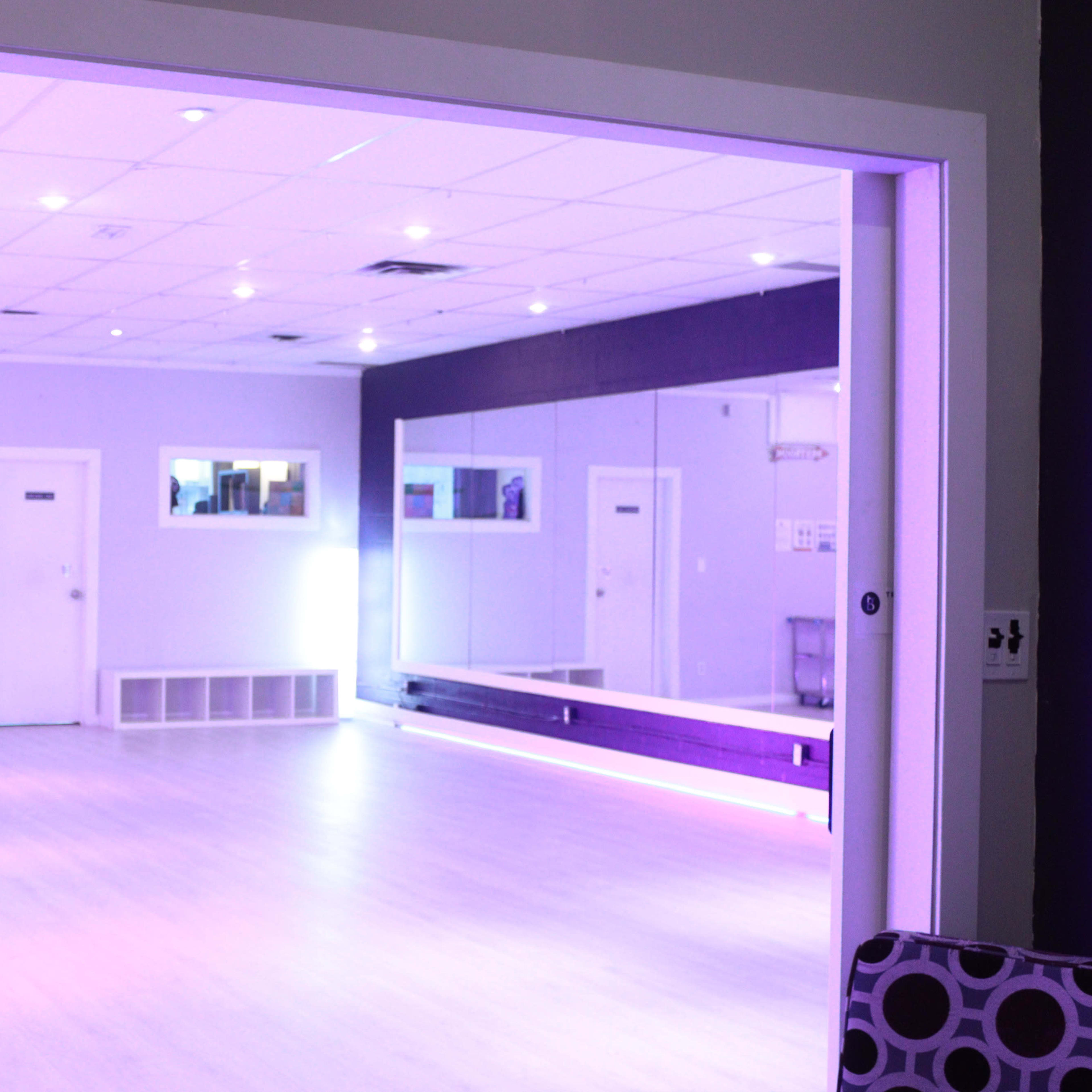 A dance studio with purple lighting features a mirrored wall and a spacious wooden floor.