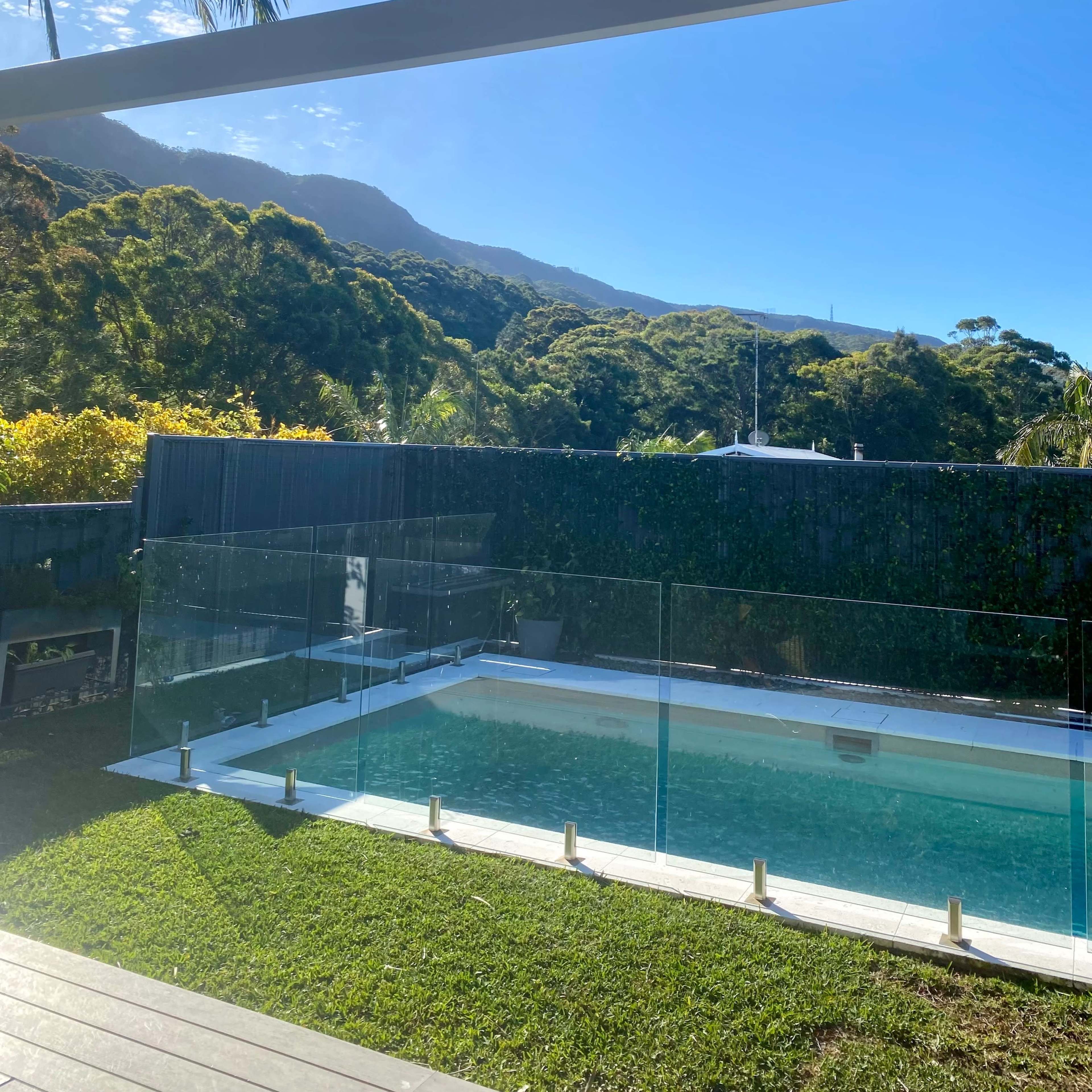 A modern swimming pool surrounded by a grassy area, with lush green mountains visible in the background under a clear blue sky.