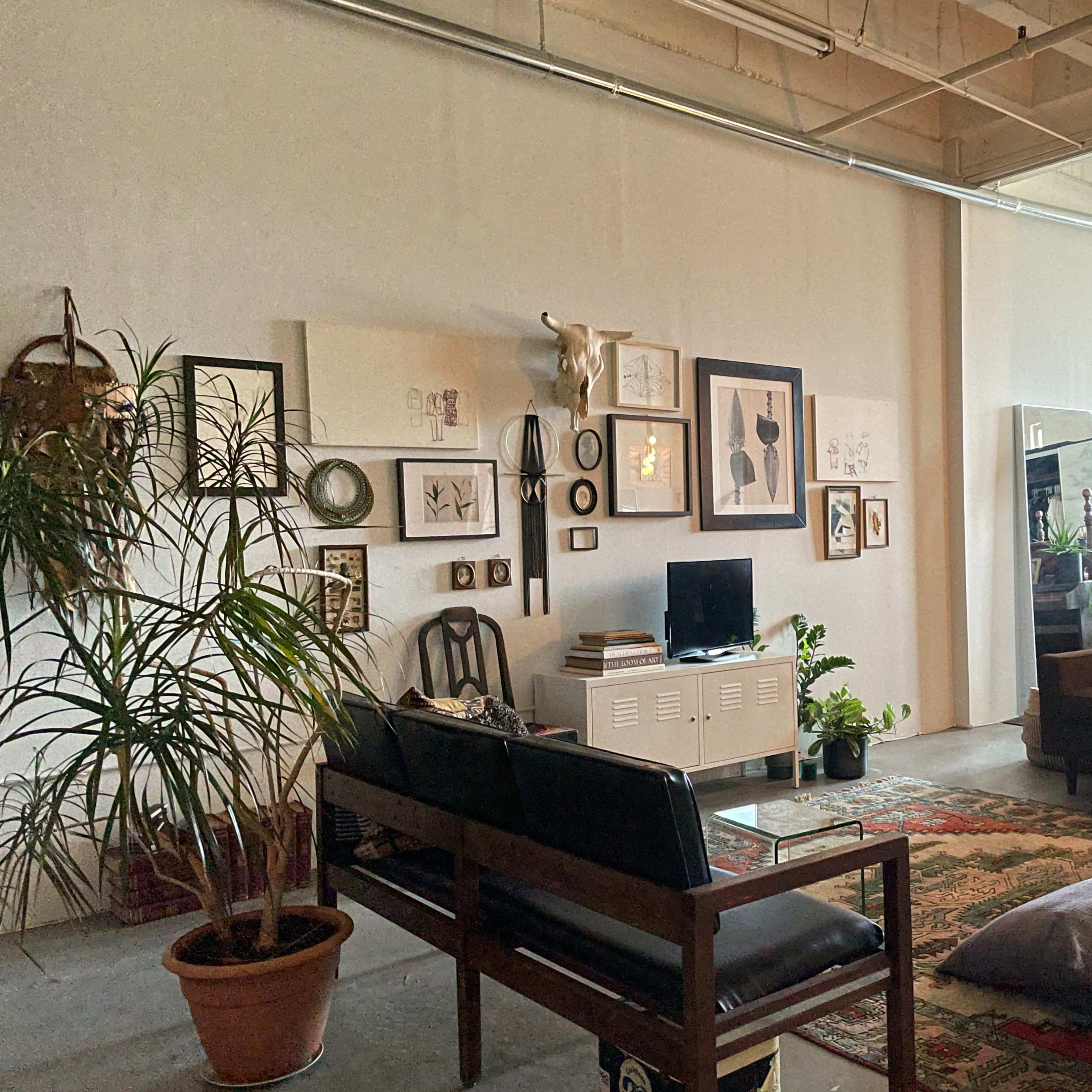 A spacious living area featuring a seating arrangement with a wooden sofa, a large rug, potted plants, wall art, and a television on a side table.