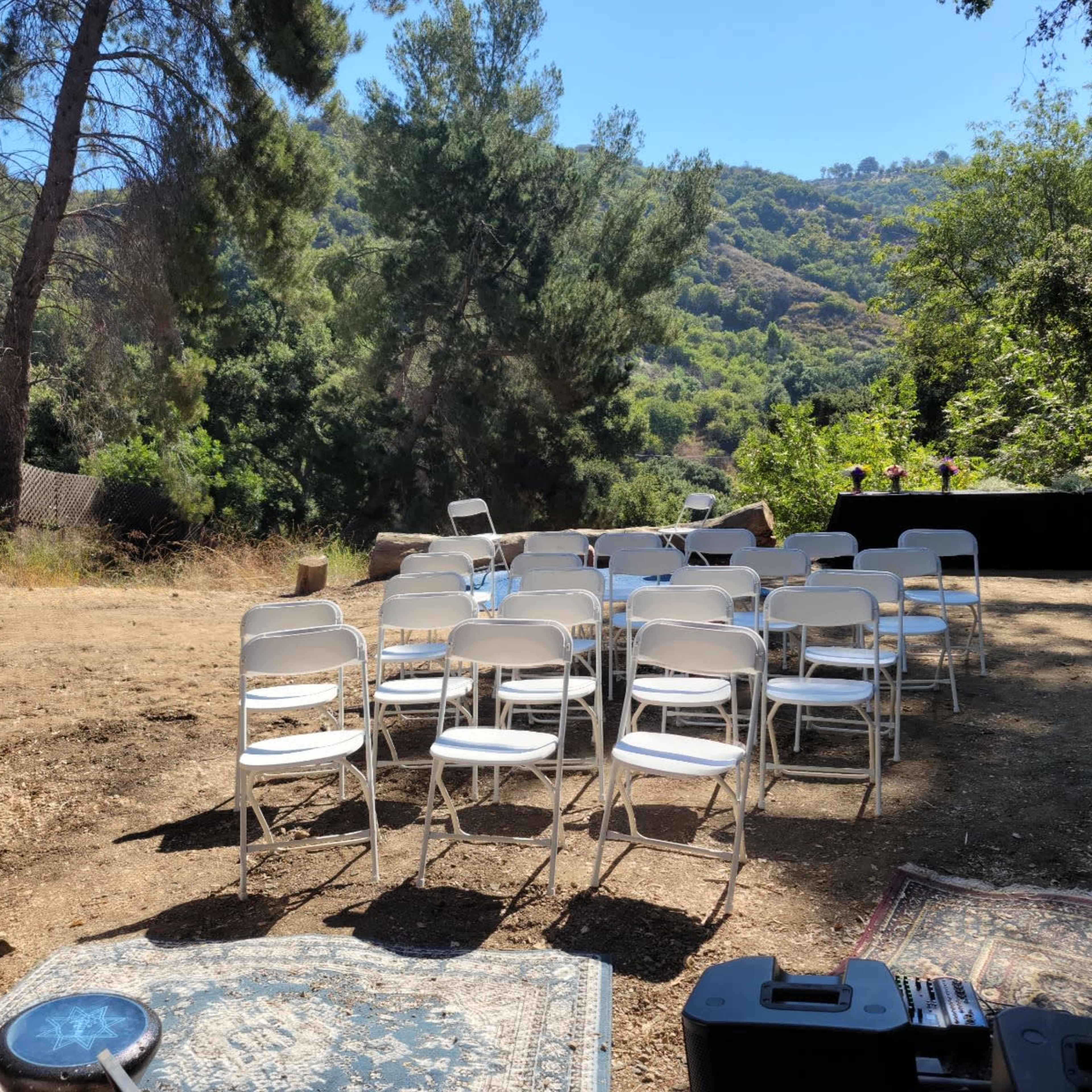 A series of white folding chairs are arranged on a dirt area surrounded by trees and hills, facing a black table set in the foreground.