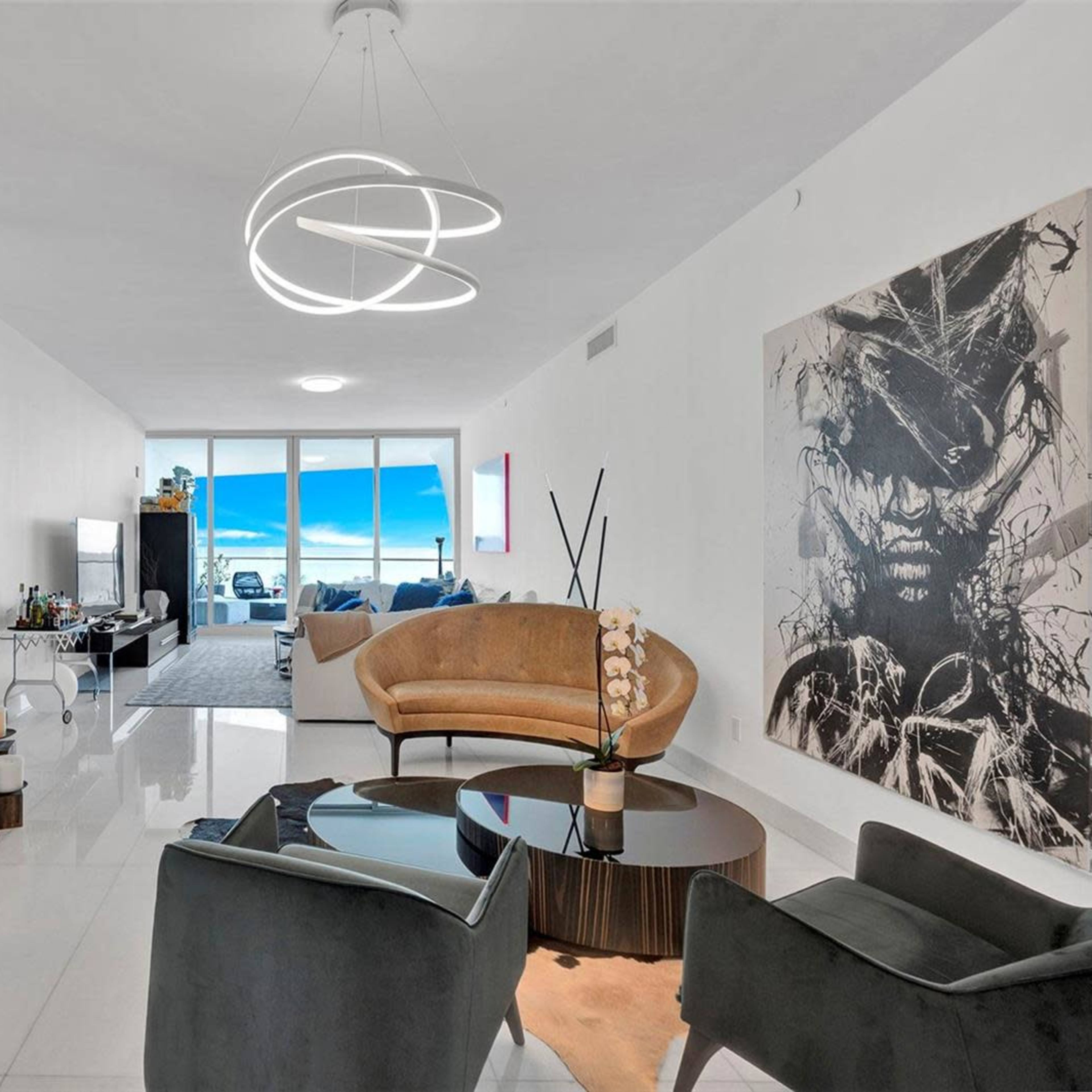 A modern living room with a large mirror, a circular light fixture, a brown sofa, and an abstract painting on the wall, overlooking a view of the outdoors through large windows.