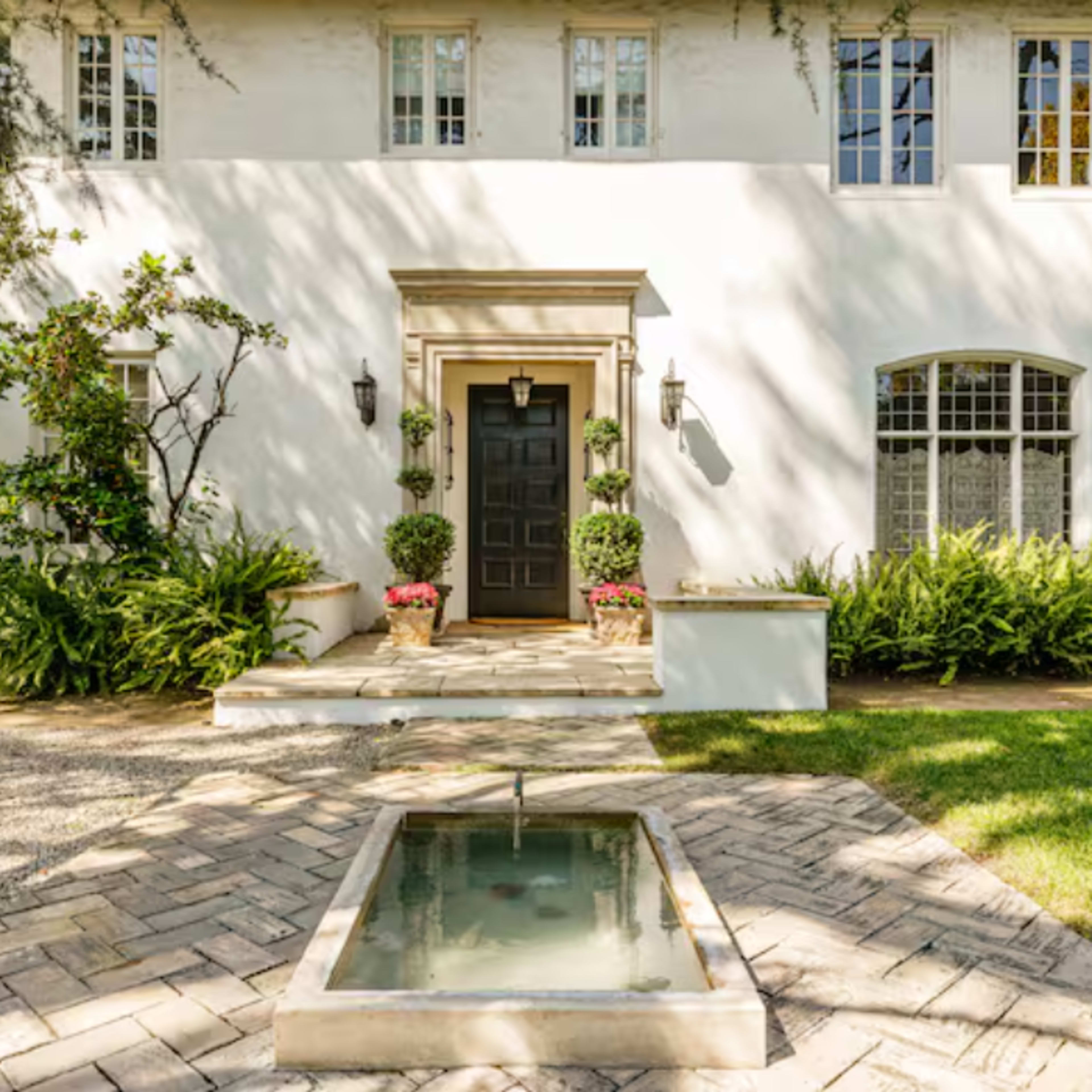 A well-maintained white house features a central black door, flanked by potted plants and a small fountain in the foreground.