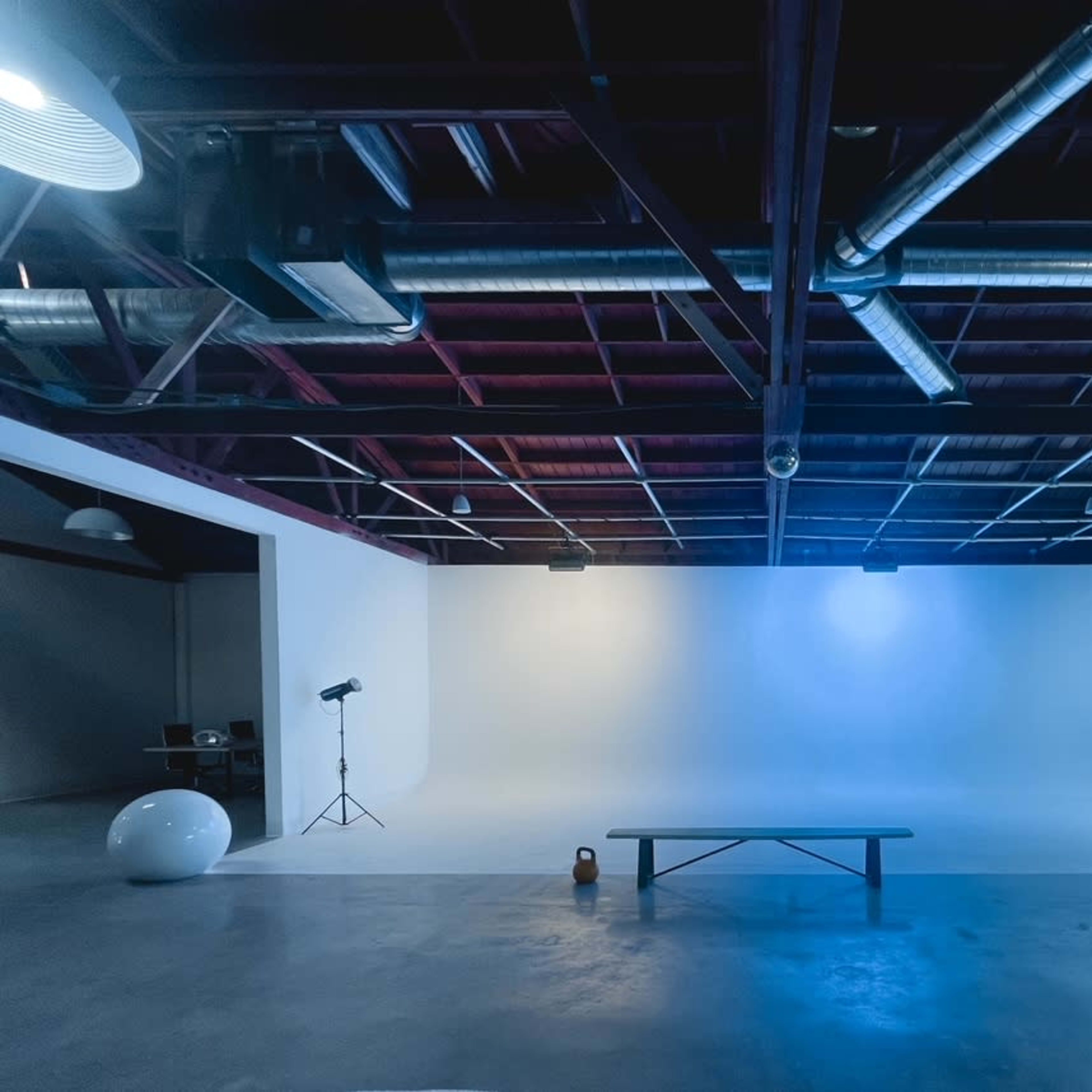 An empty studio space features a smooth white backdrop, a wooden bench, a round object, and various lighting equipment suspended from the ceiling.