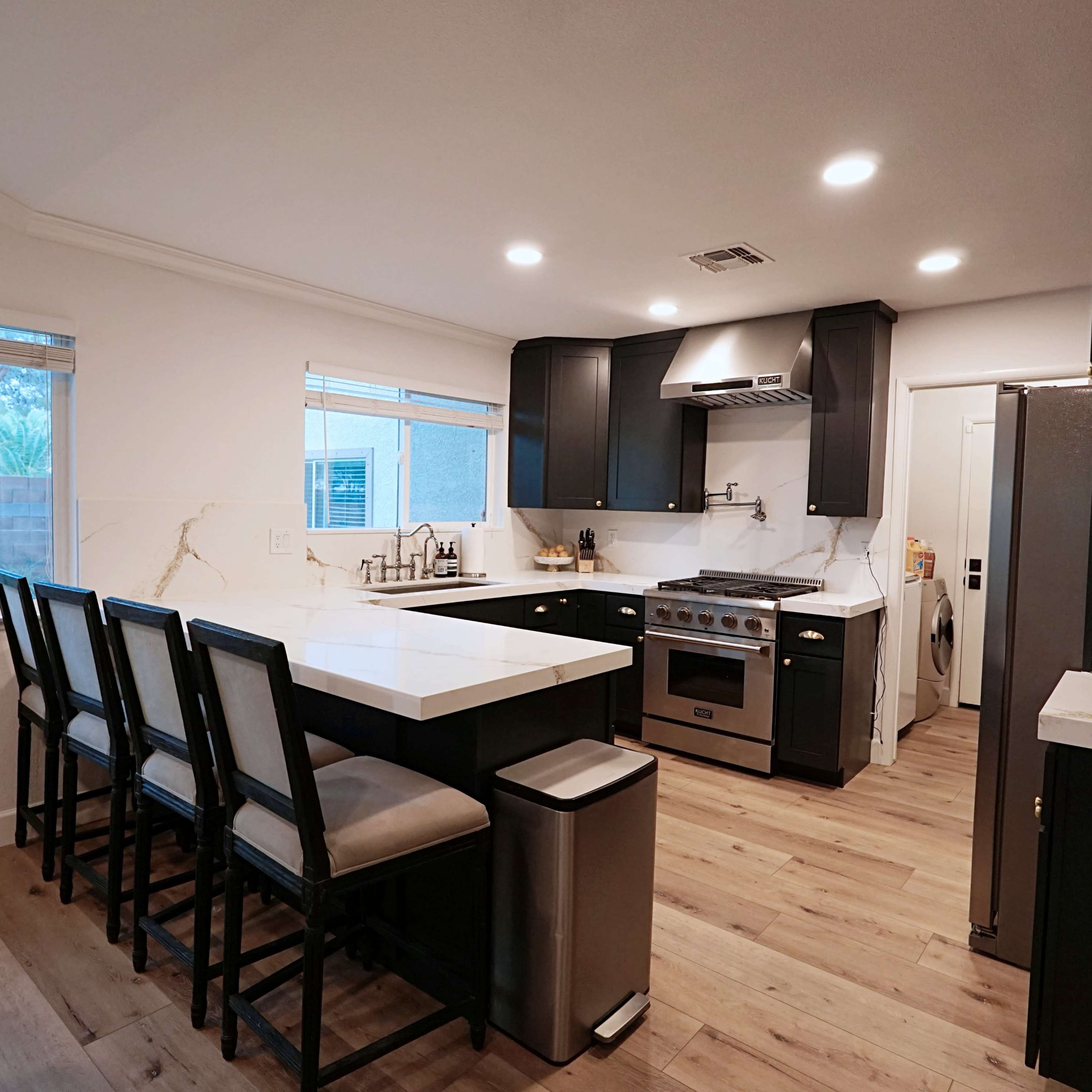 A modern kitchen features dark cabinetry, a large island with seating, stainless steel appliances, and ample natural light from windows.