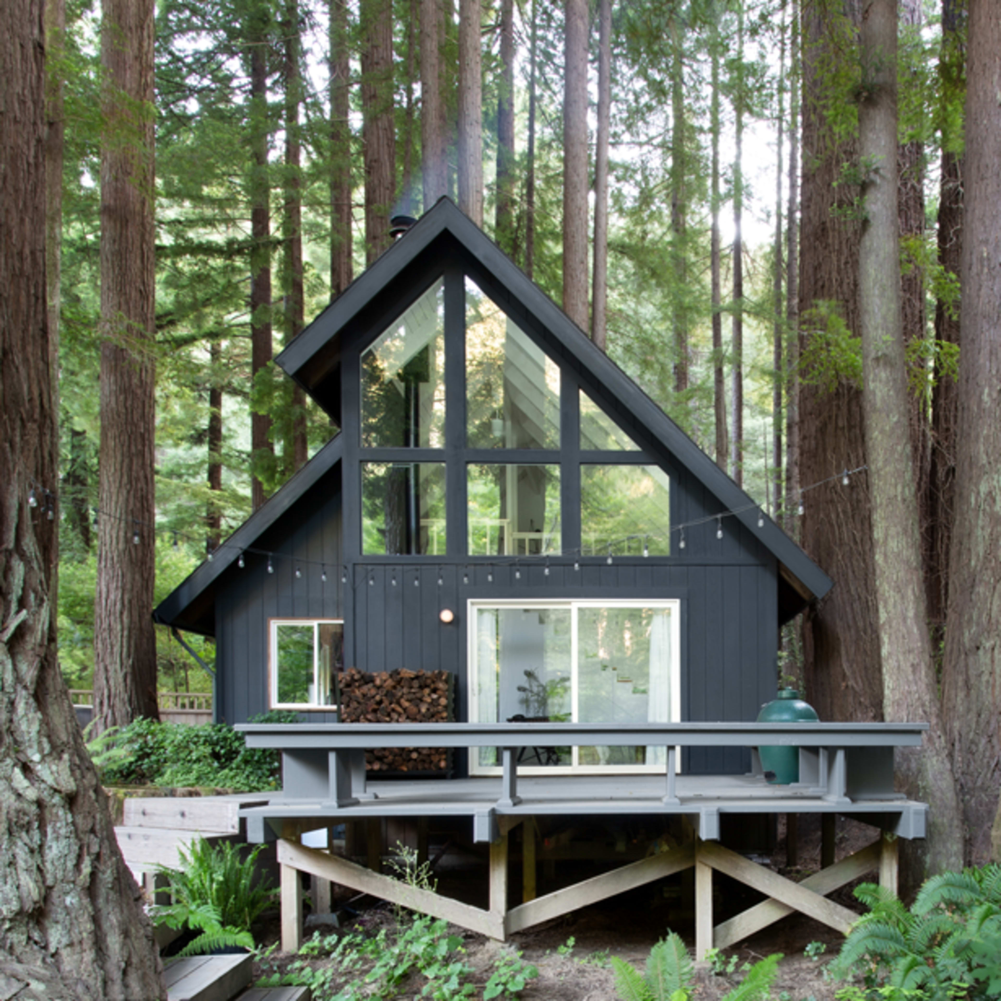 A black wooden cabin with large windows stands among tall redwood trees, featuring a deck with a table and stacked firewood.