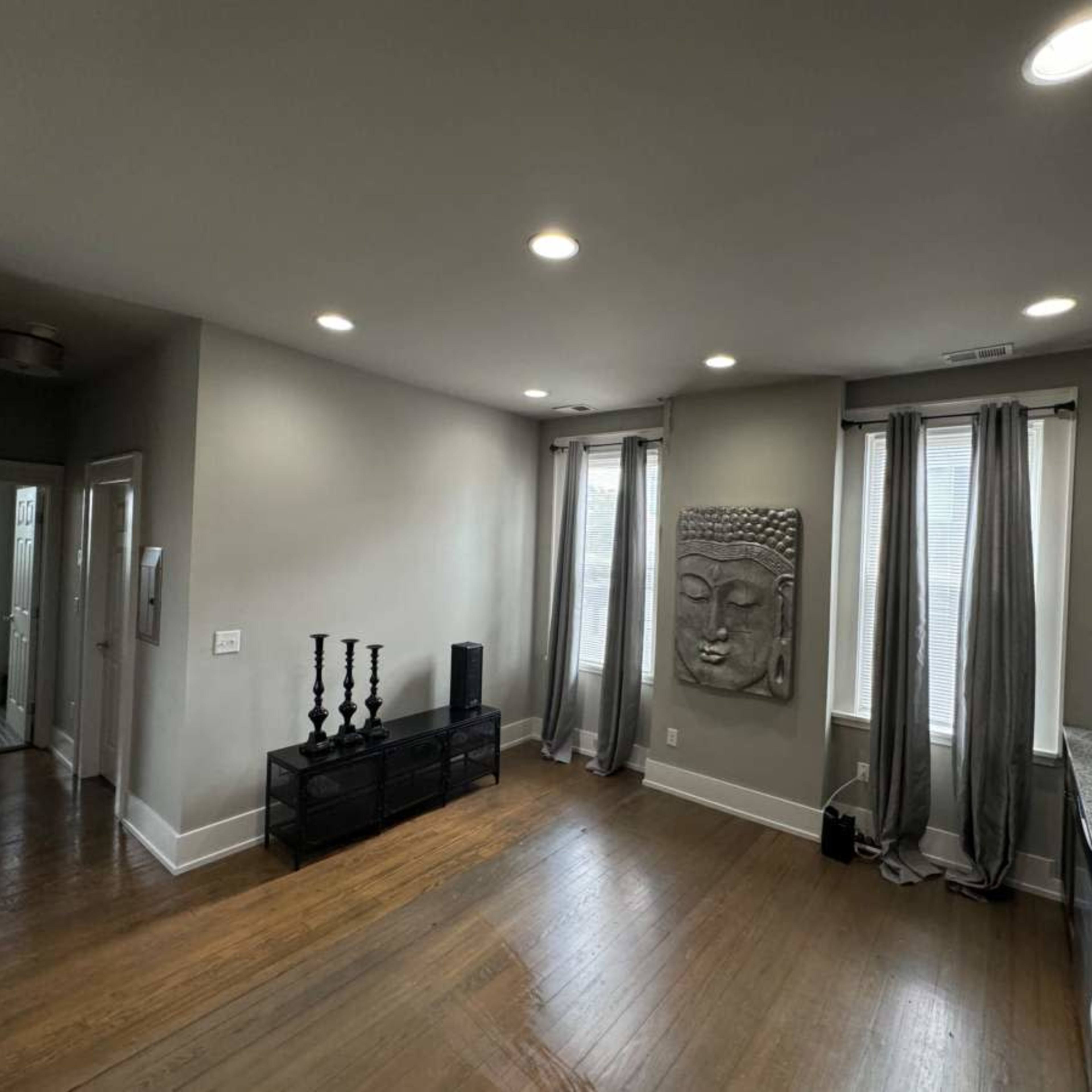 The room features wooden flooring, gray walls, a large Buddha wall sculpture, and a kitchen area with dark cabinets and a countertop.