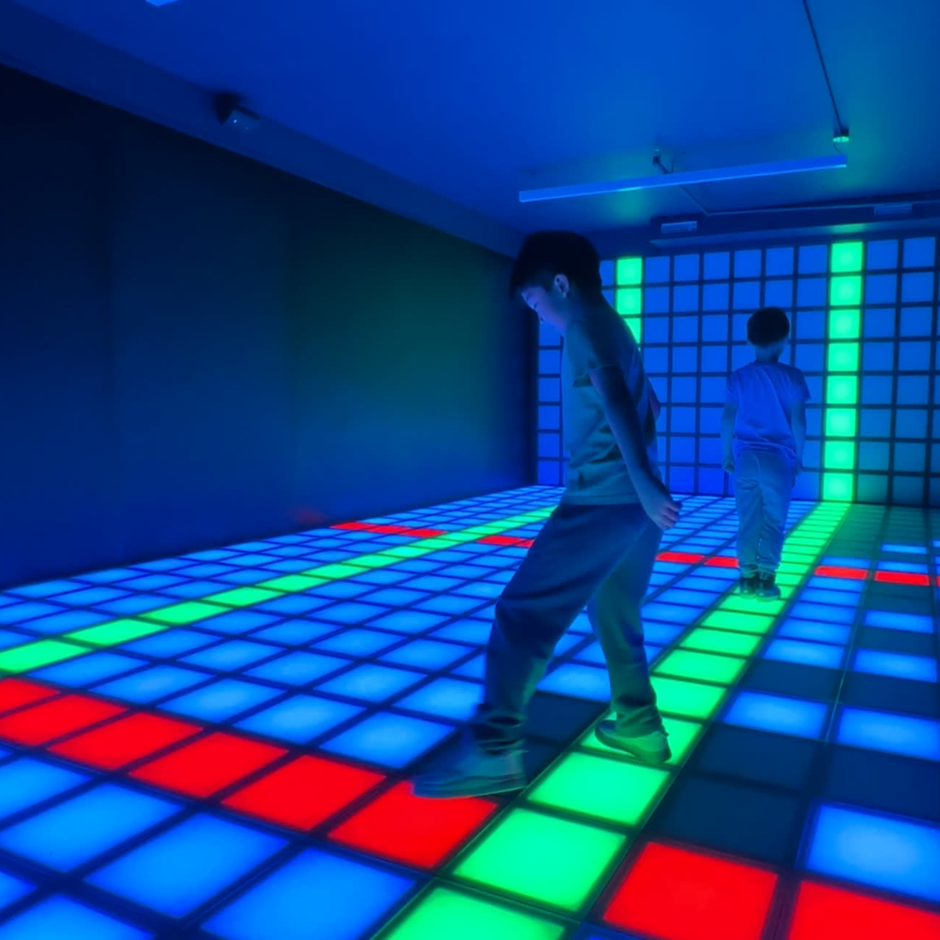 Two individuals walk on a brightly lit grid floor with blue, green, and red squares in a dimly lit room.