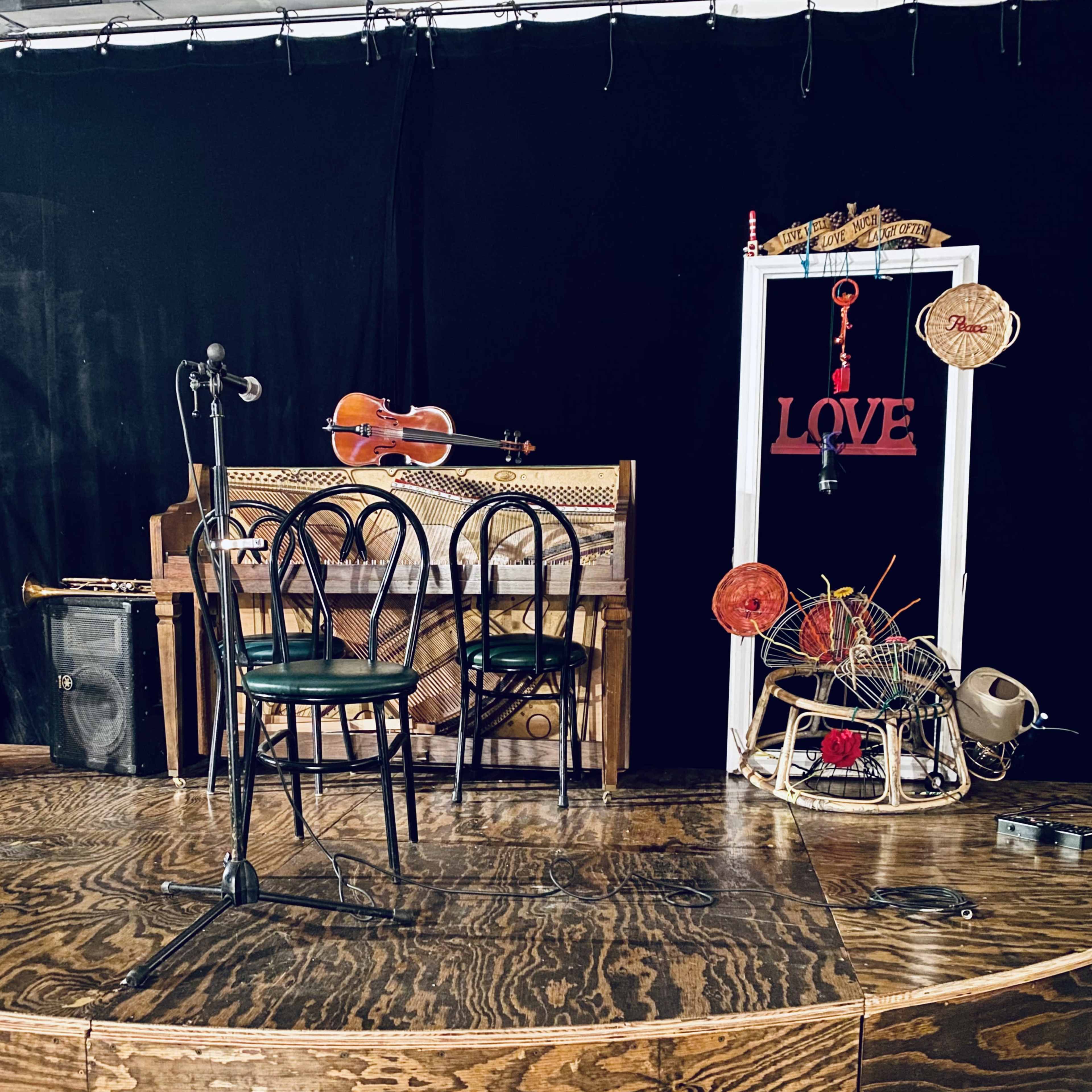 A small stage with two chairs, a microphone, a double bass, a violin, a piano, and various decorative items, all set against a black backdrop.