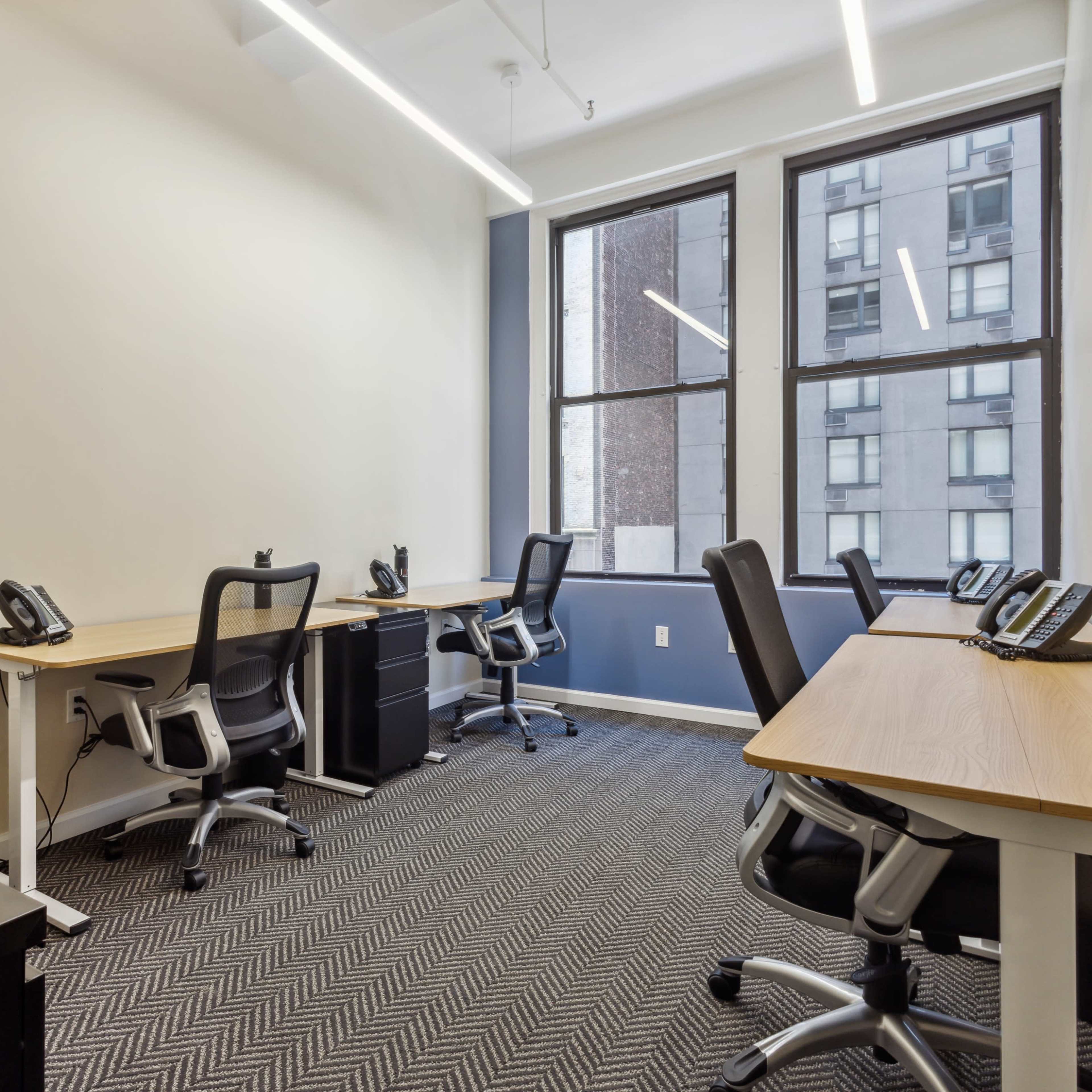 A modern office space with several desks and chairs, each equipped with a telephone, and large windows providing natural light.