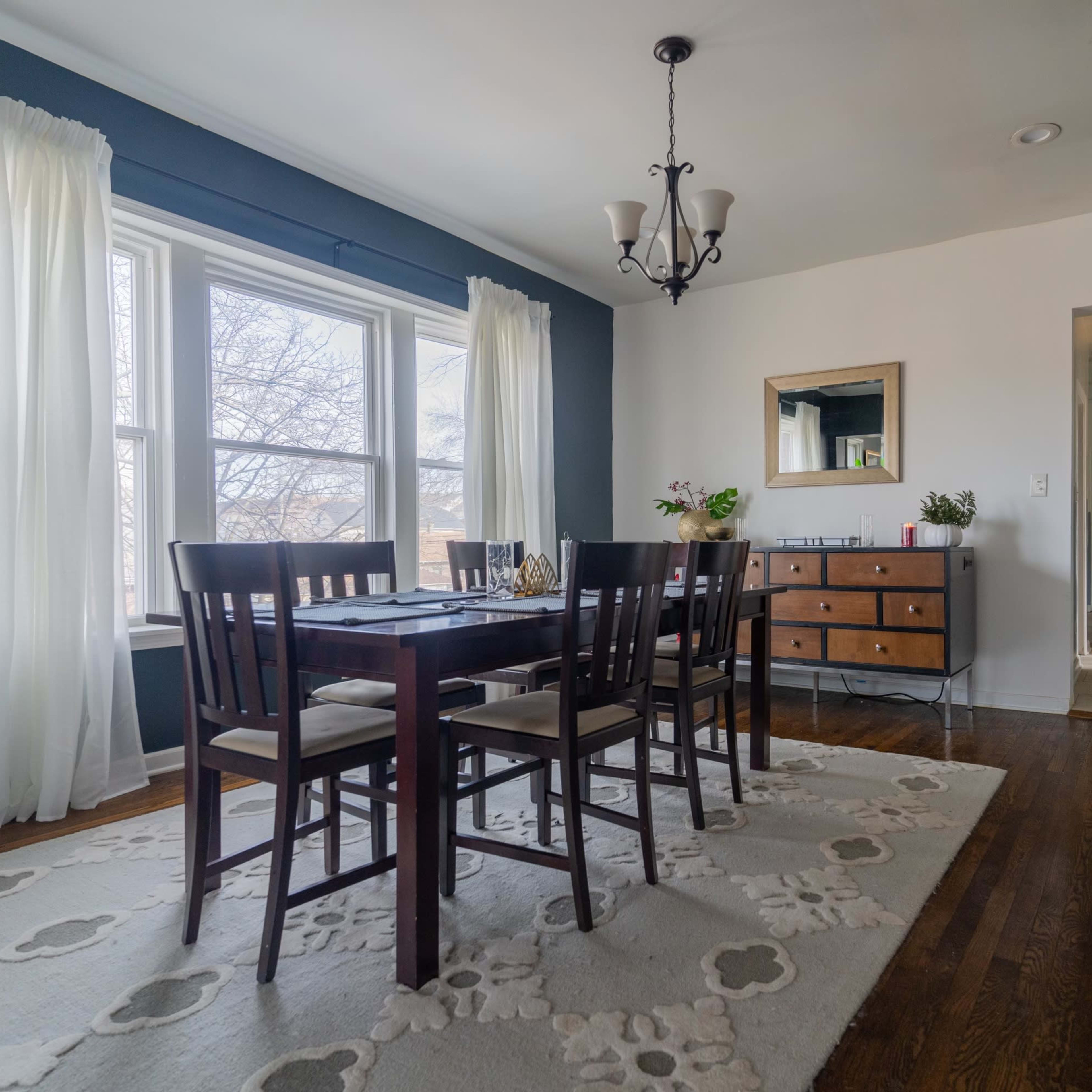 A dining room features a wooden table surrounded by chairs, large windows with sheer curtains, and a sideboard, with an adjoining hallway visible.