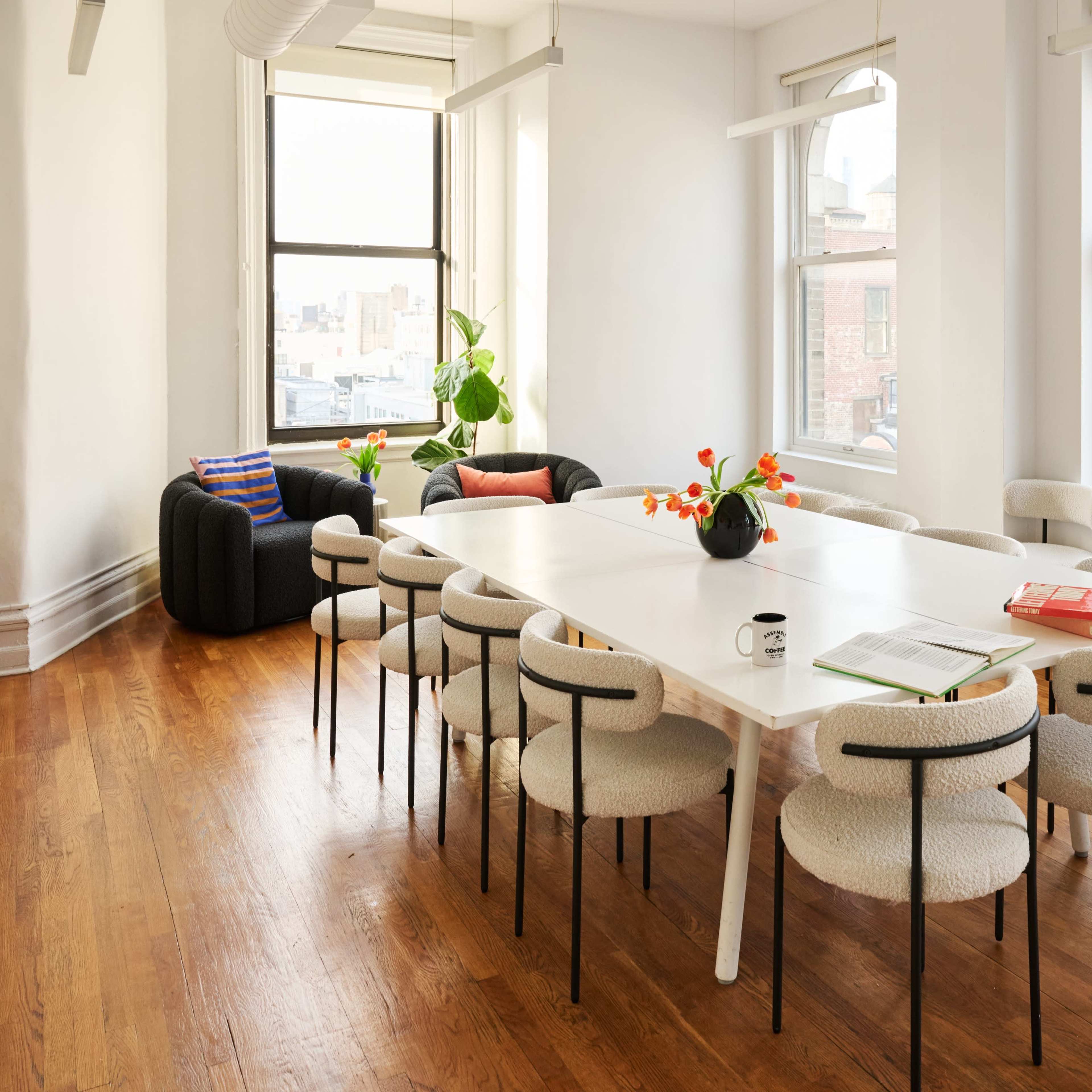A bright, modern meeting room features a long white table surrounded by ten chairs, with large windows allowing natural light and a plant in the corner.
