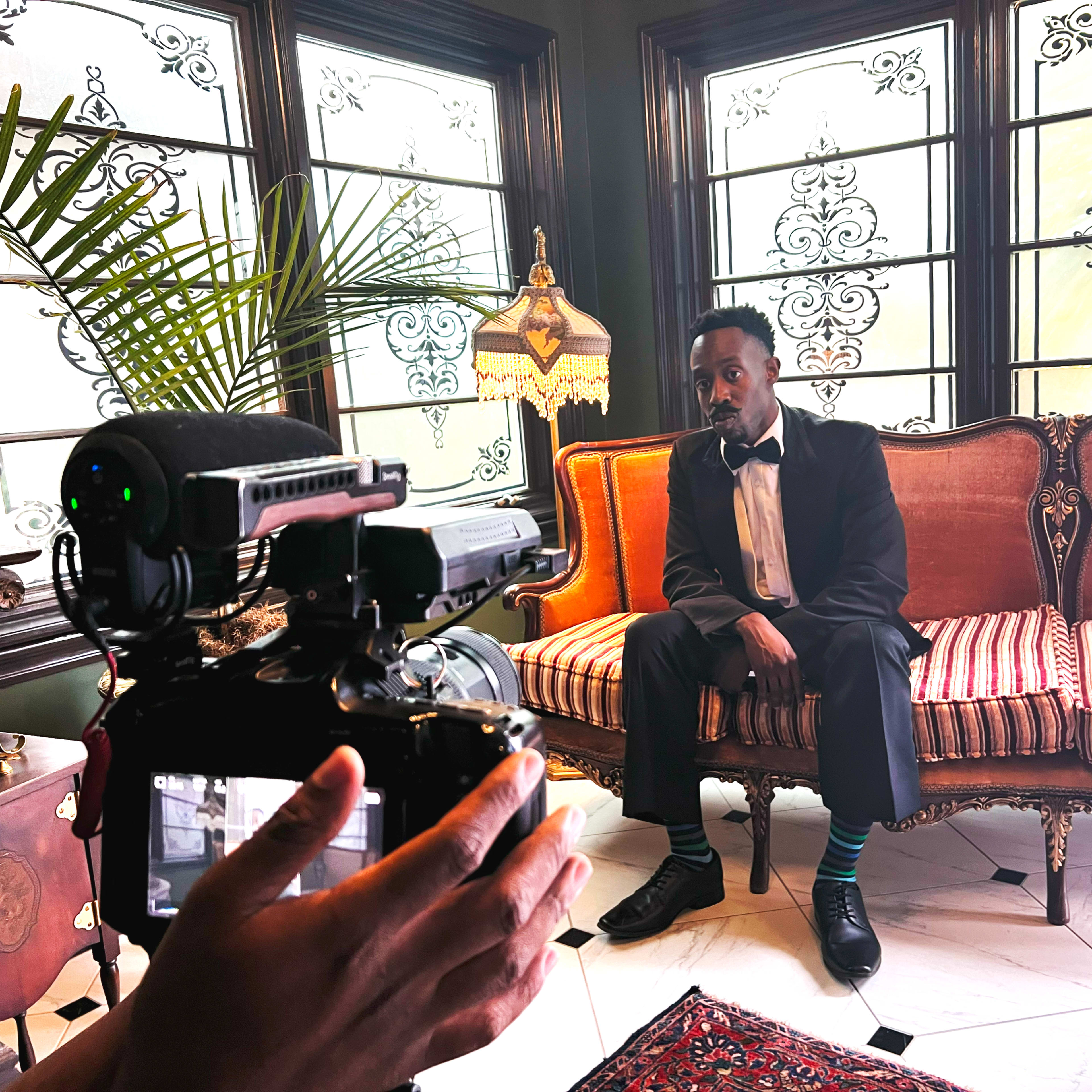 A person dressed in formal attire sits on a striped sofa while a camera is directed at them in a well-decorated room.