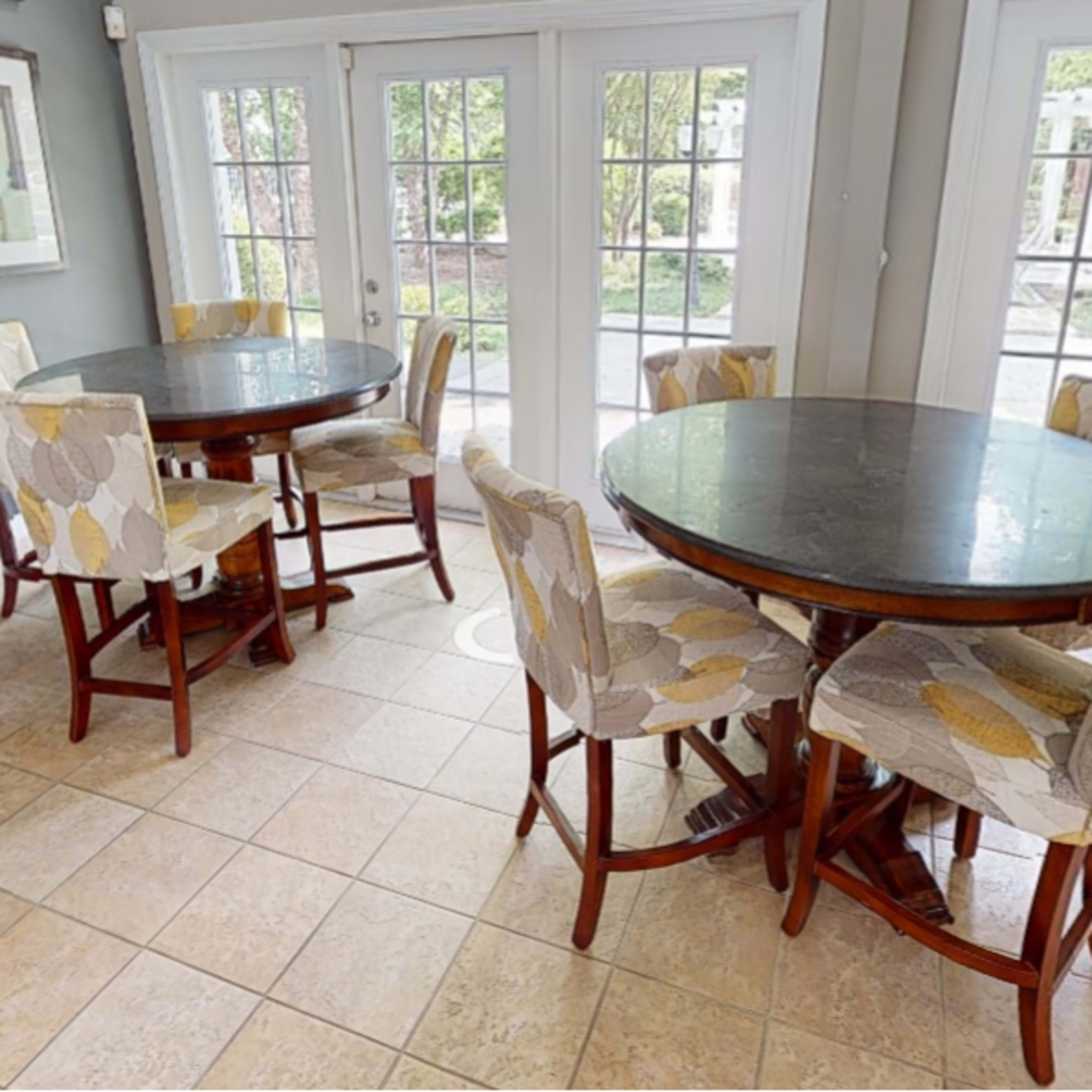 A bright room with two round dining tables, each surrounded by six upholstered chairs, and large windows allowing natural light to enter.