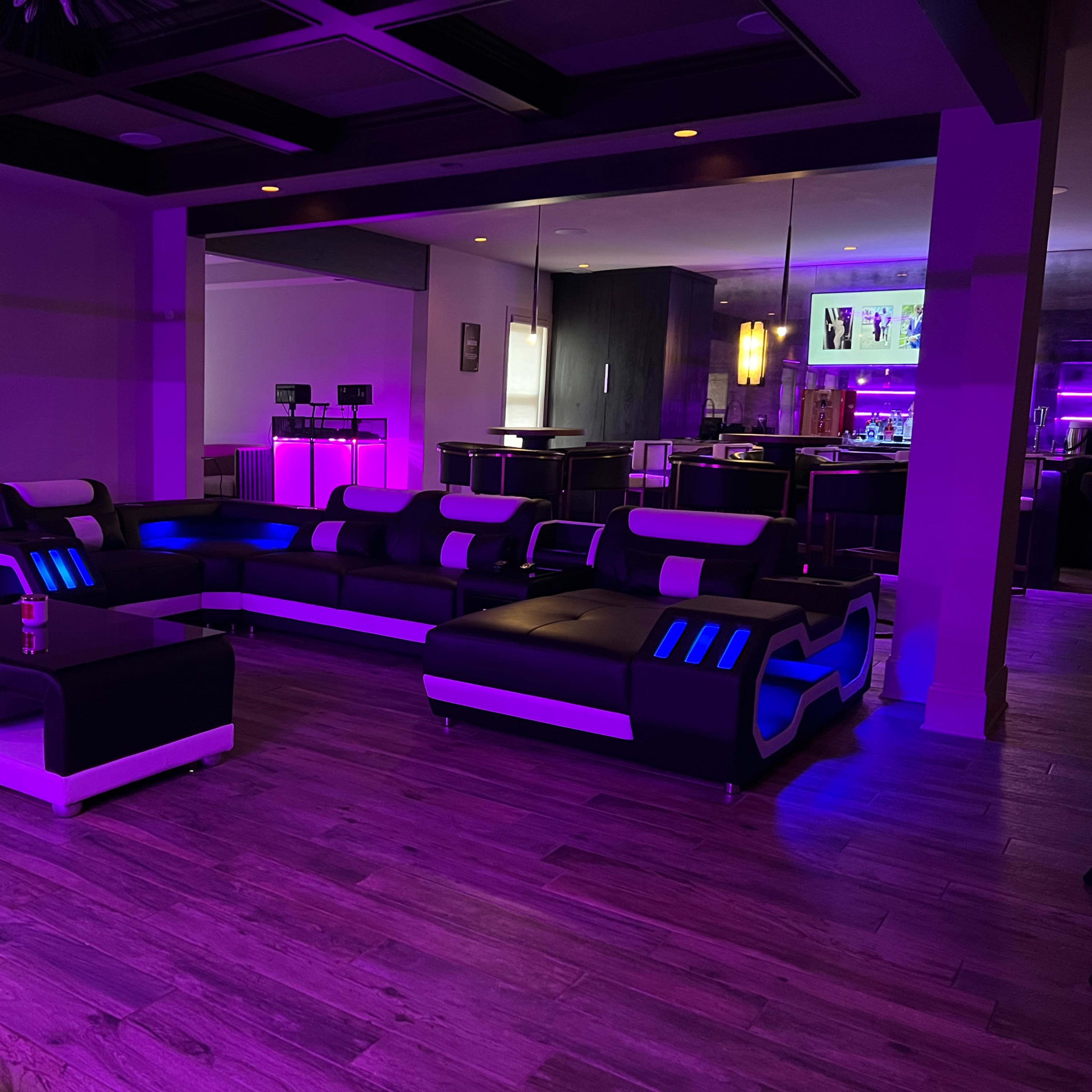 A modern living room featuring a black and white sectional sofa with blue LED lighting and a matching coffee table, set against a backdrop of purple accent lighting.