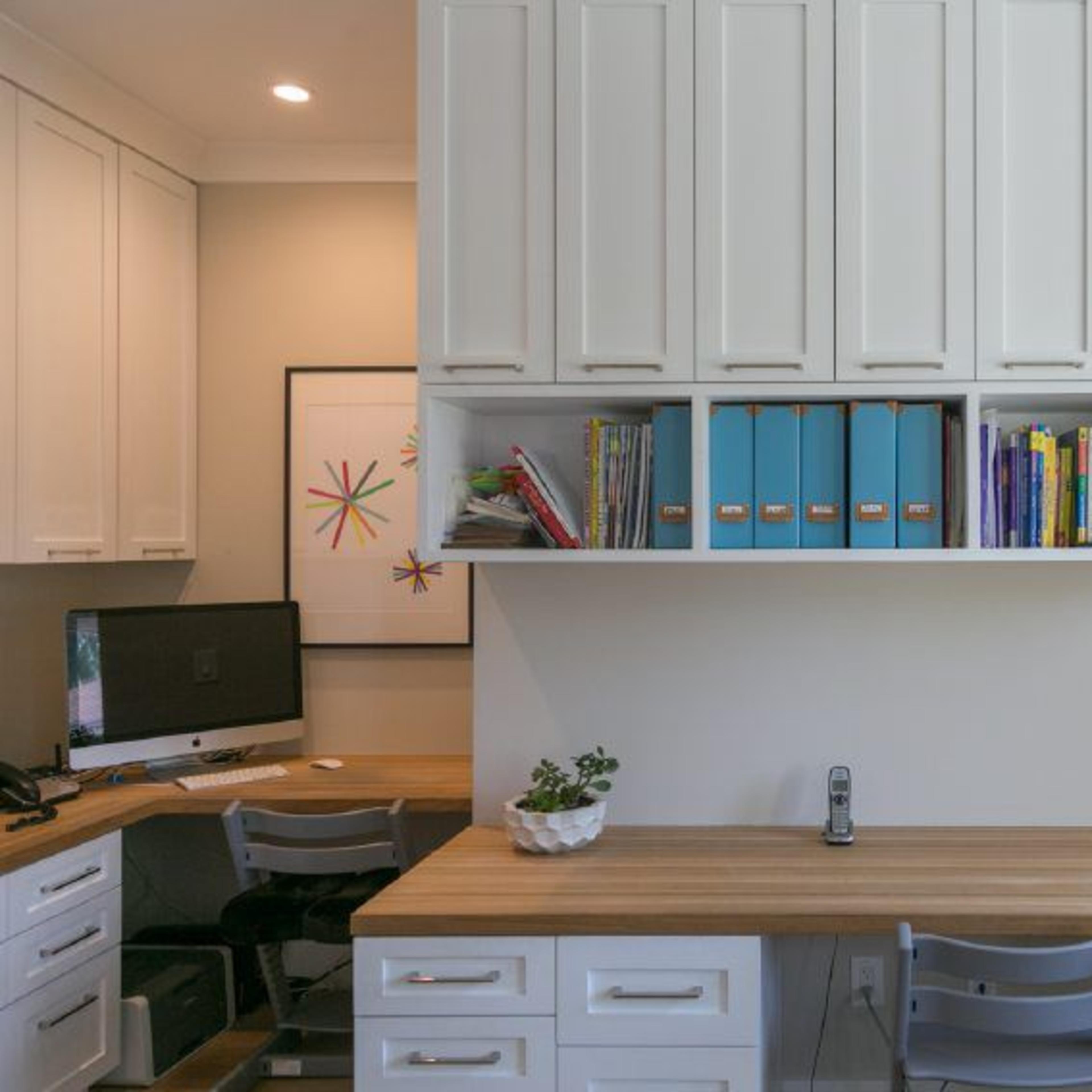 A modern office space featuring a wooden desk, white cabinetry, colorful books and folders on shelves, and a computer station in the background.