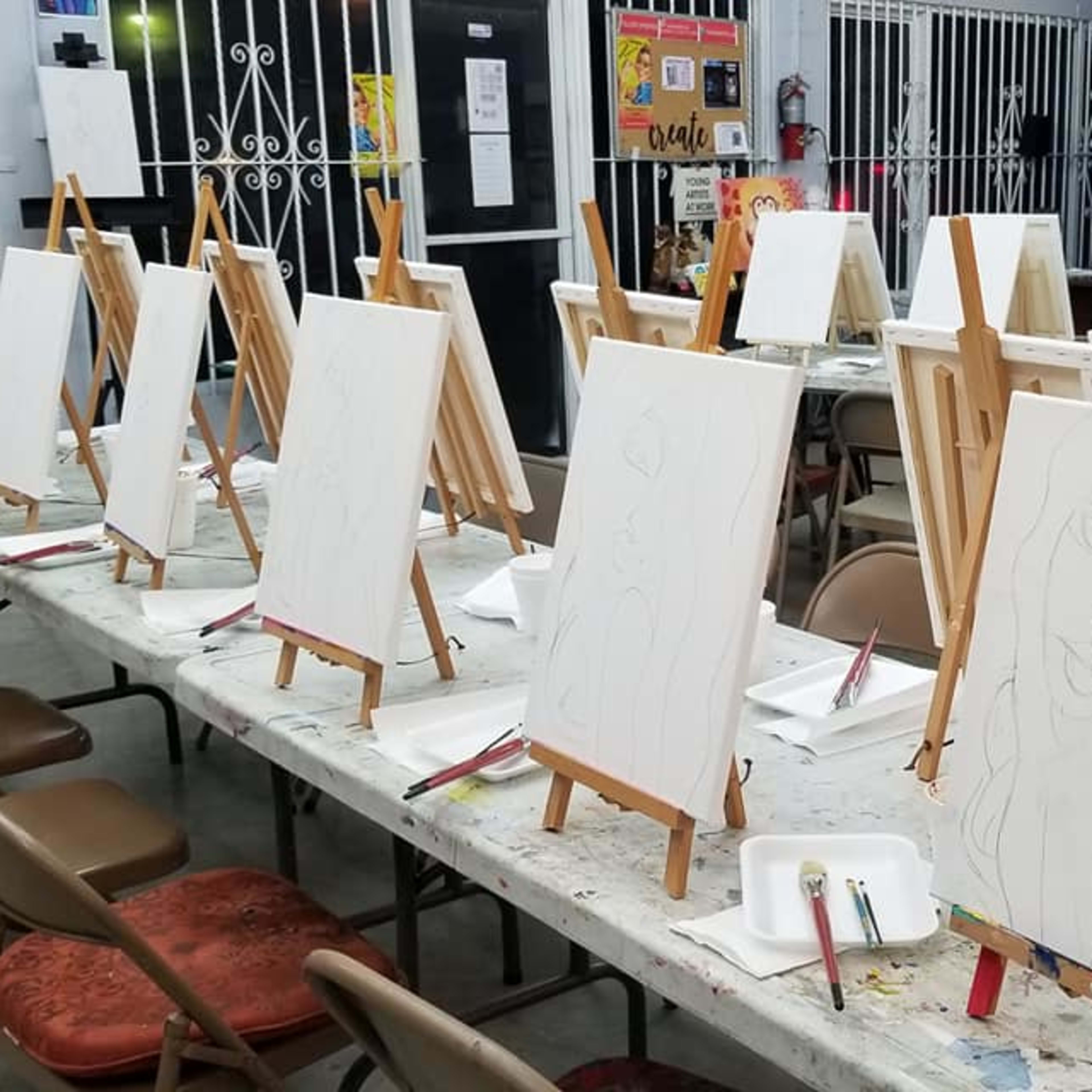 A row of blank canvases on easels set up on tables in an art studio.