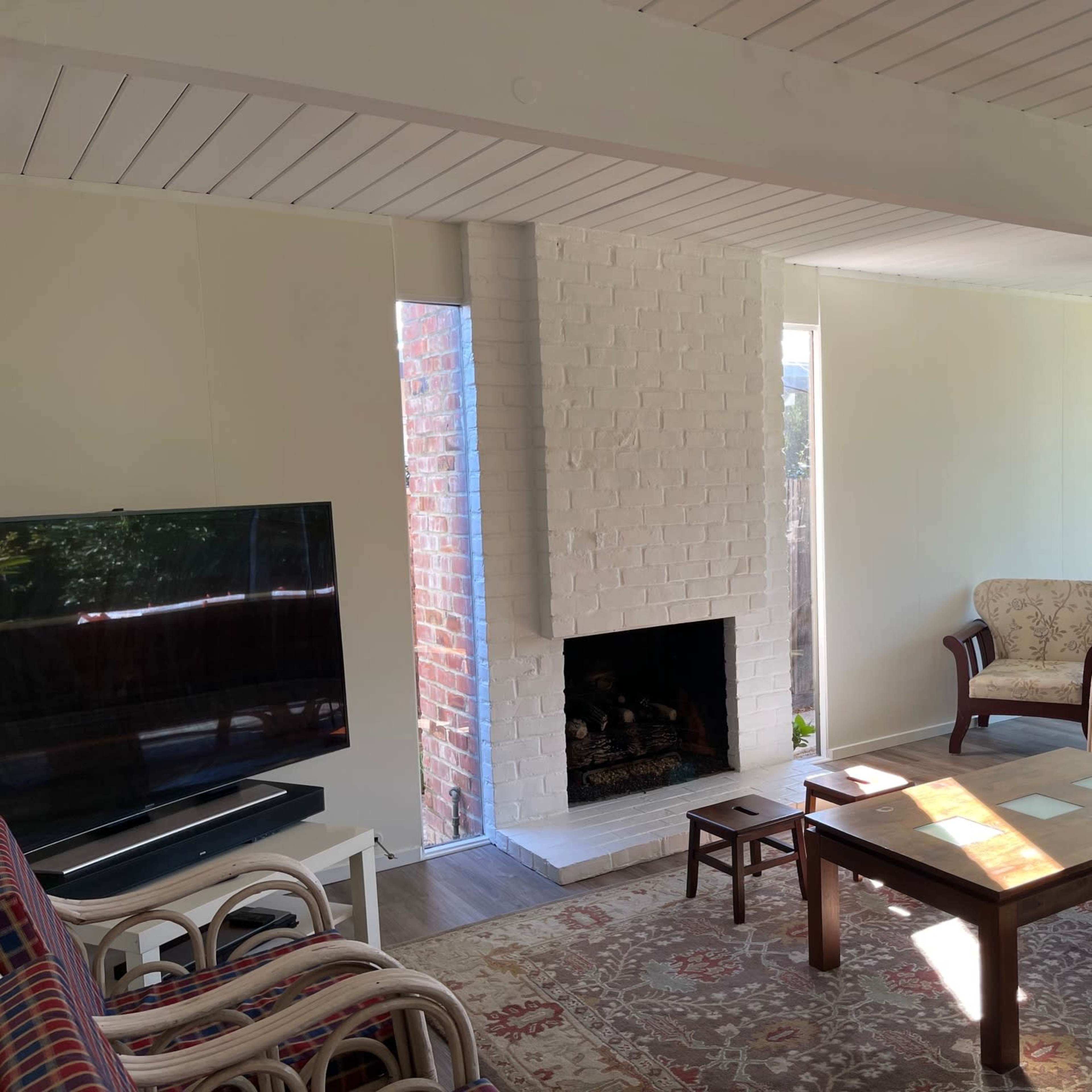 A living room with a white brick fireplace, a television, a patterned sofa, a wooden coffee table, and large windows allowing natural light in.