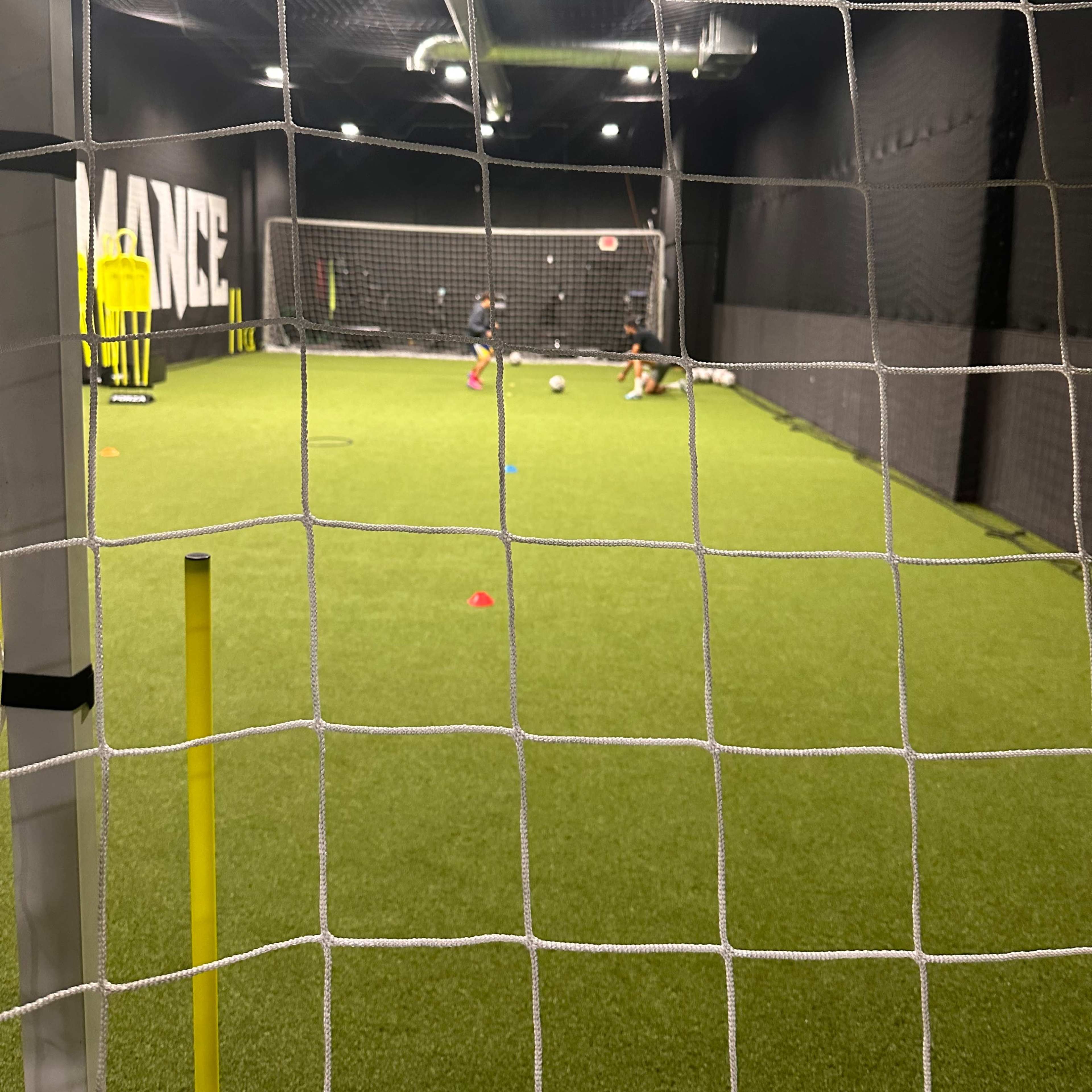 Two players practice soccer drills on a synthetic grass field inside a training facility, with goals and training equipment visible in the background.