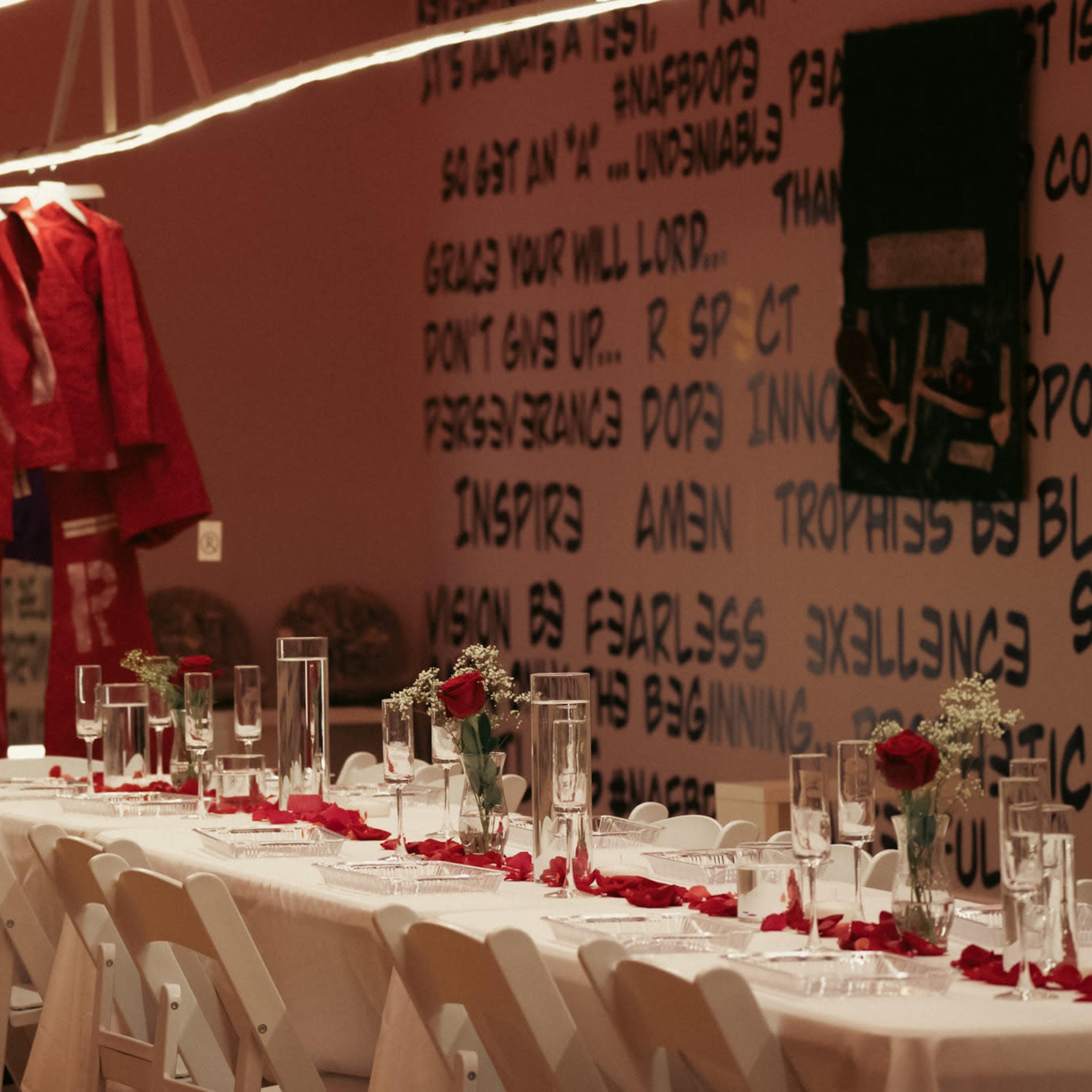 A long dining table set with glasses and rose petals is positioned under hanging clothing displays in a designed room.