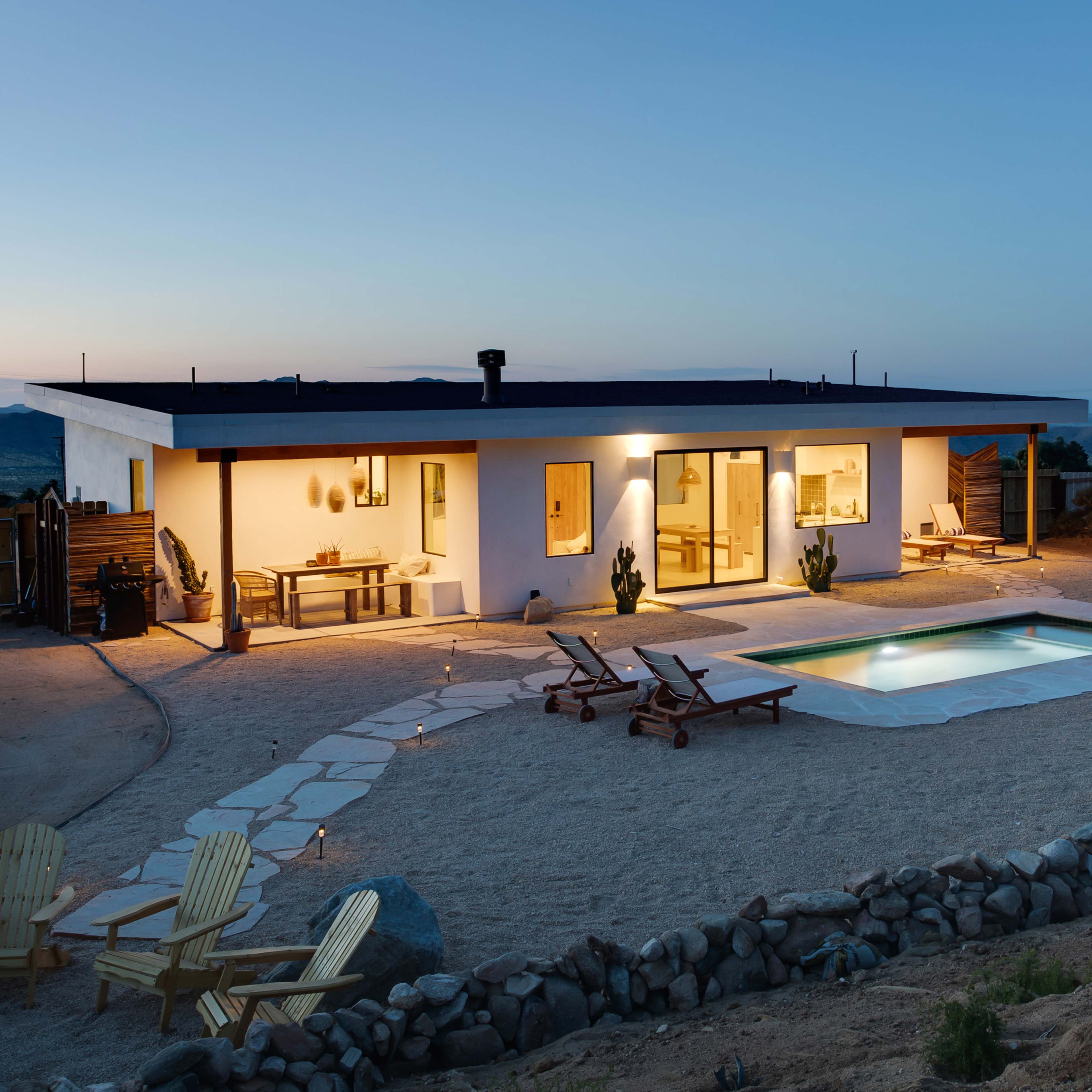 A modern house with a pool is illuminated at dusk, surrounded by a sandy area and desert landscaping.