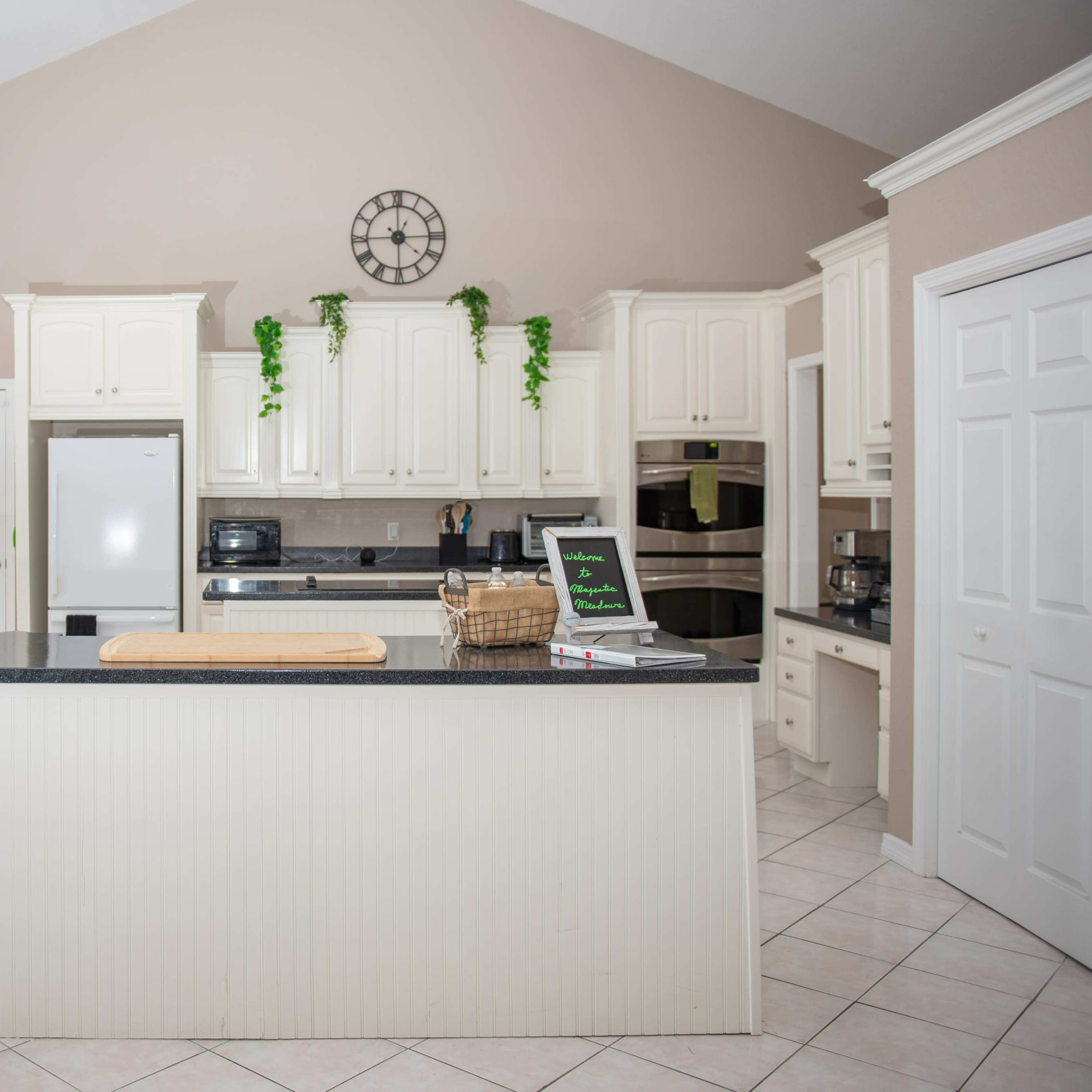 The kitchen features white cabinetry, a large dark countertop island, and stainless steel appliances with green plants decorating the shelves.