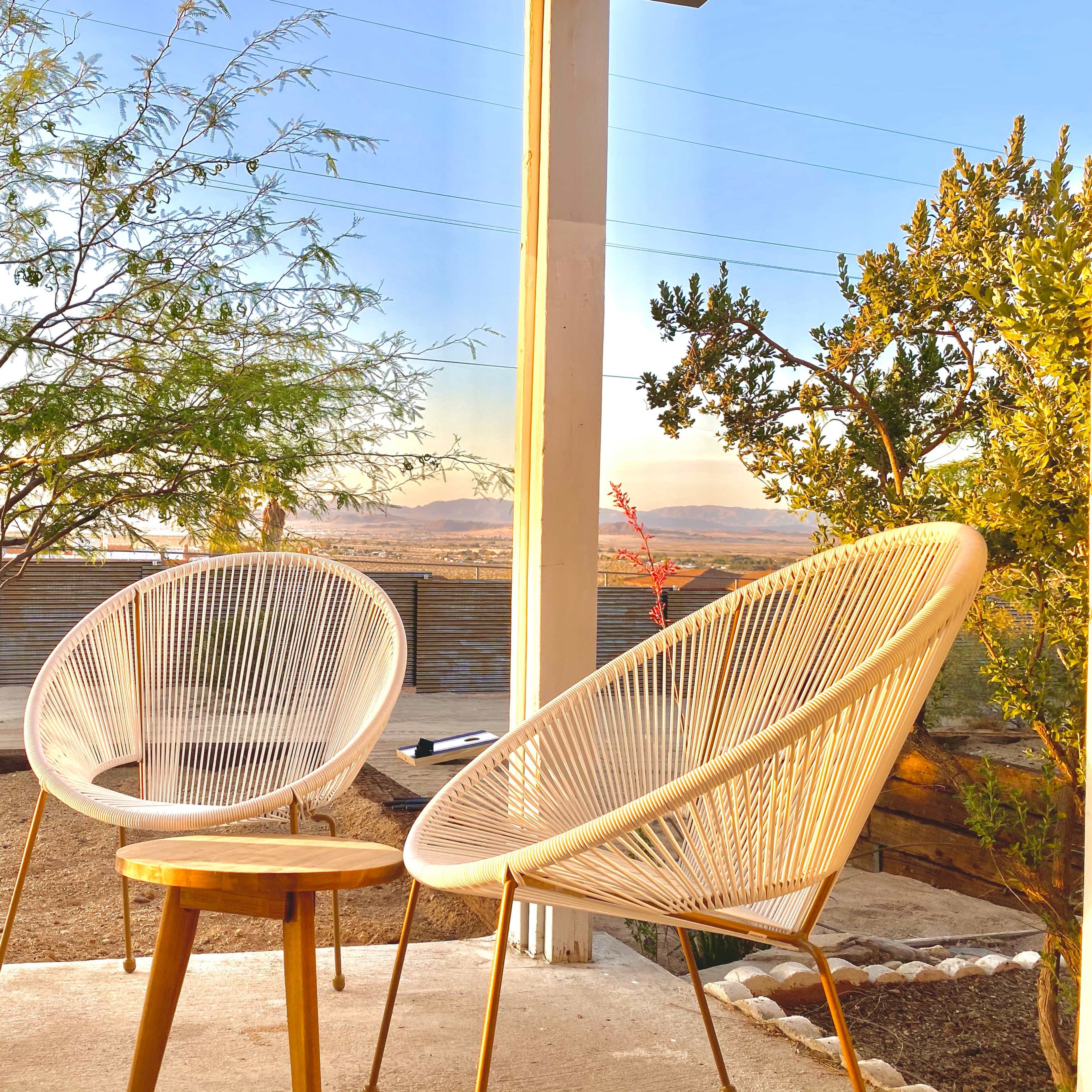 Two white, woven chairs and a small round table sit on a patio, overlooking a desert landscape with a tree and low shrubs.