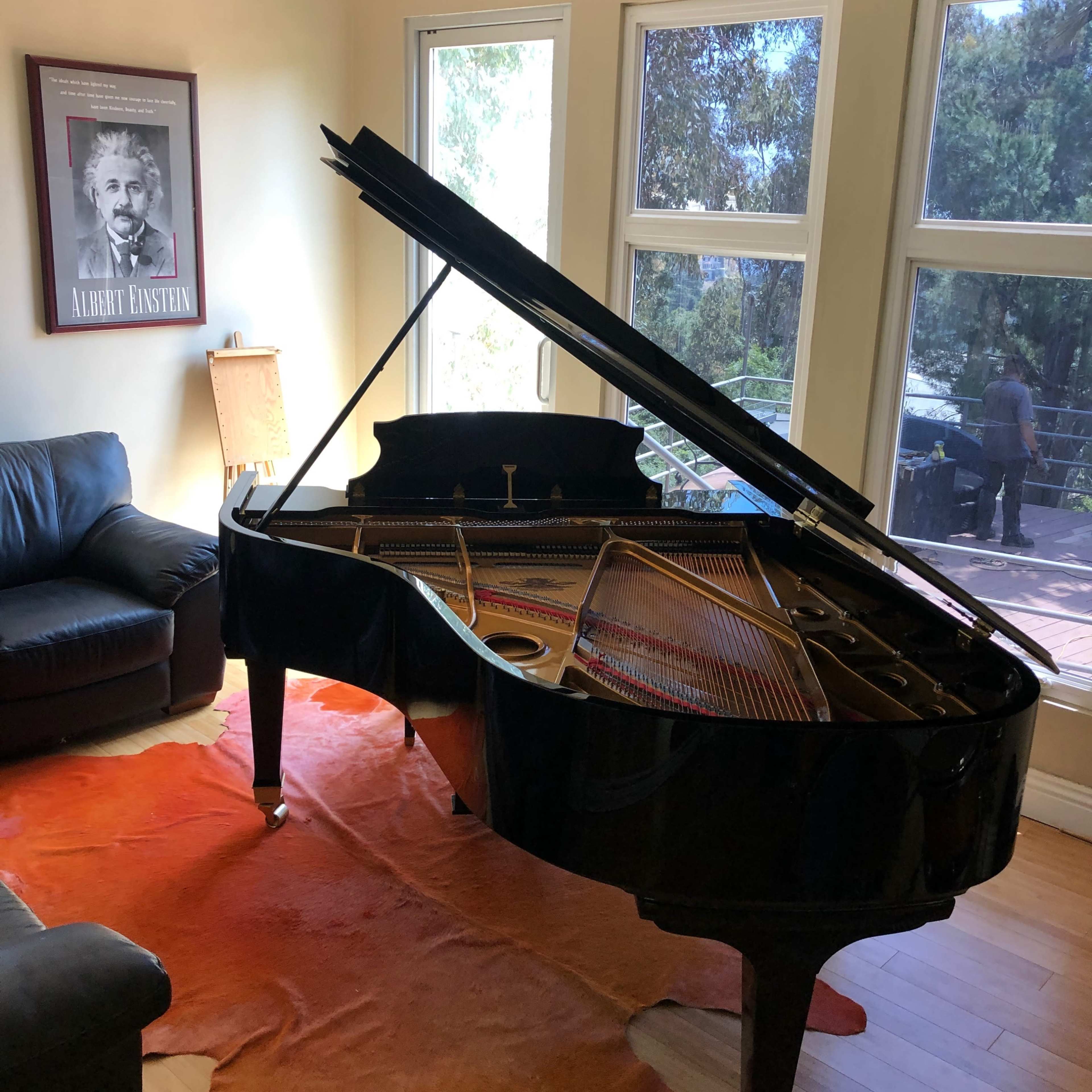 A grand piano with its lid open sits on a brown rug in a room with large windows, overlooking a patio area.