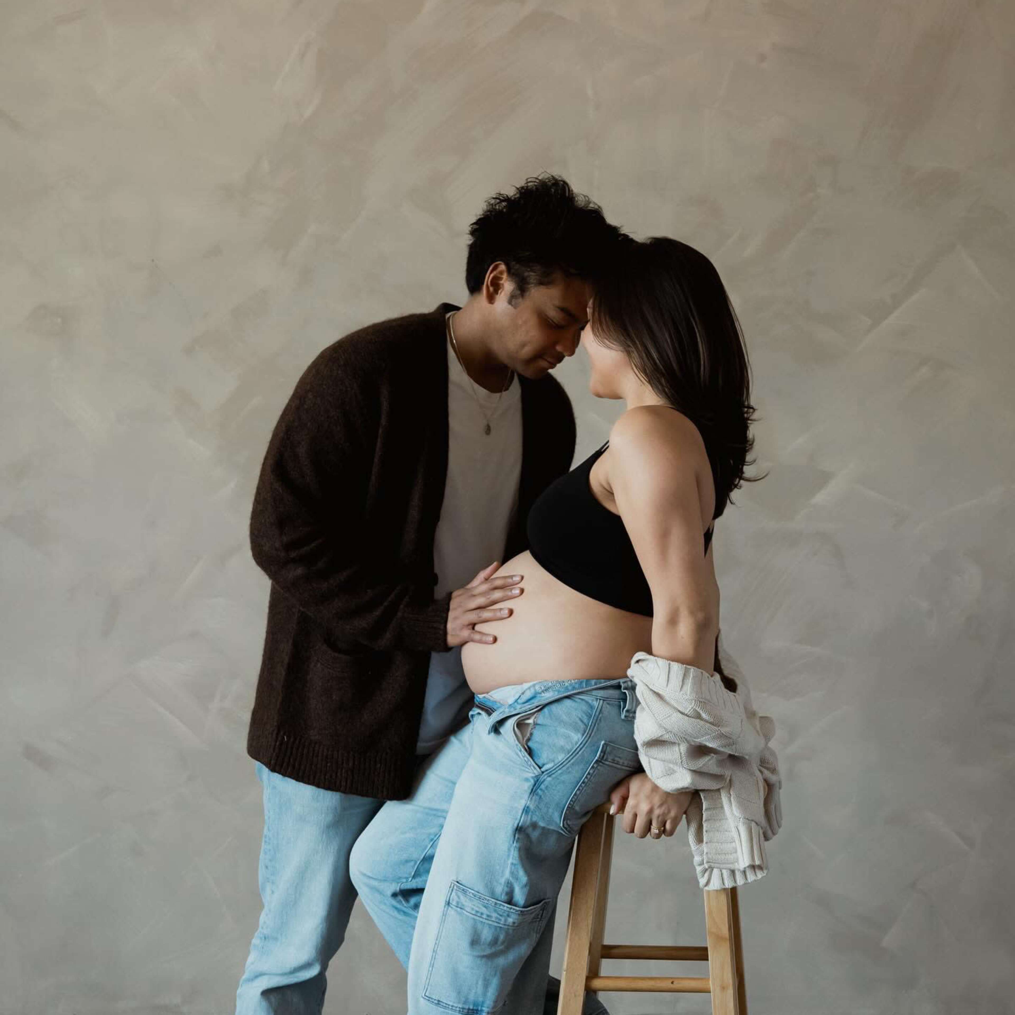 A couple stands close together, with the woman resting her hands on her pregnant belly while leaning against a wooden stool.