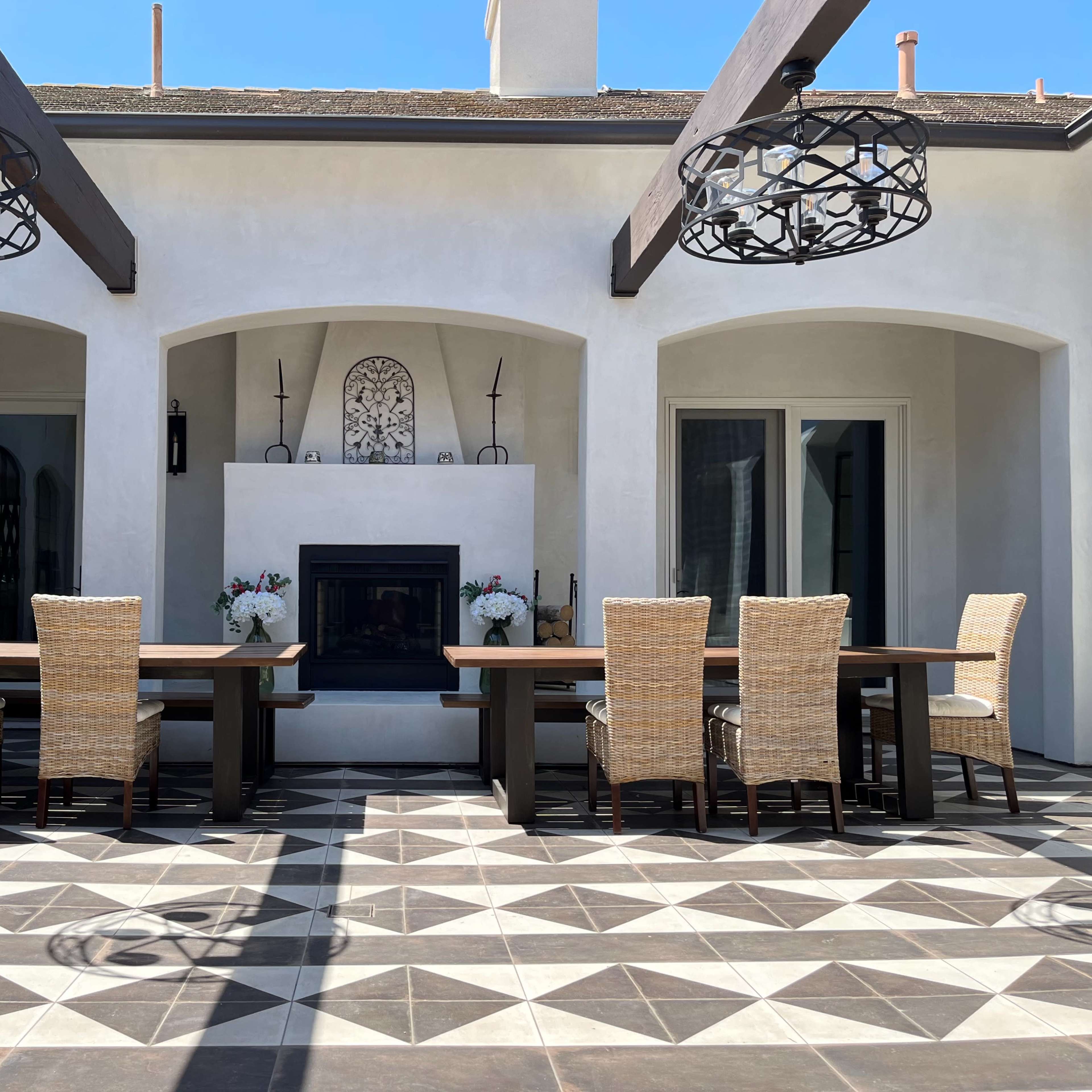 An outdoor dining area with two long tables surrounded by wicker chairs, a fireplace in the center, and large arching doorways.