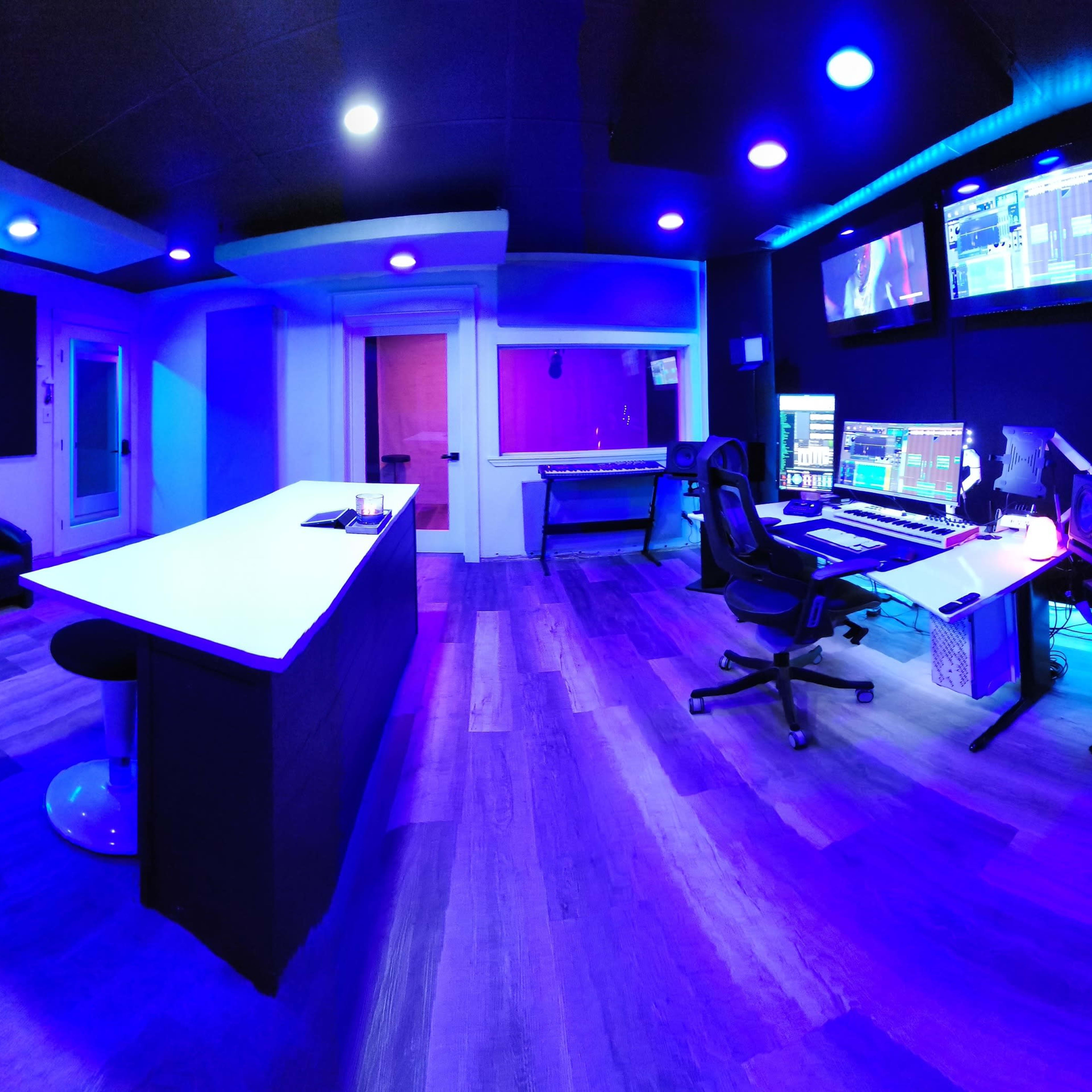 A modern recording studio with a long counter, a large desk equipped with audio equipment, and blue ambient lighting.