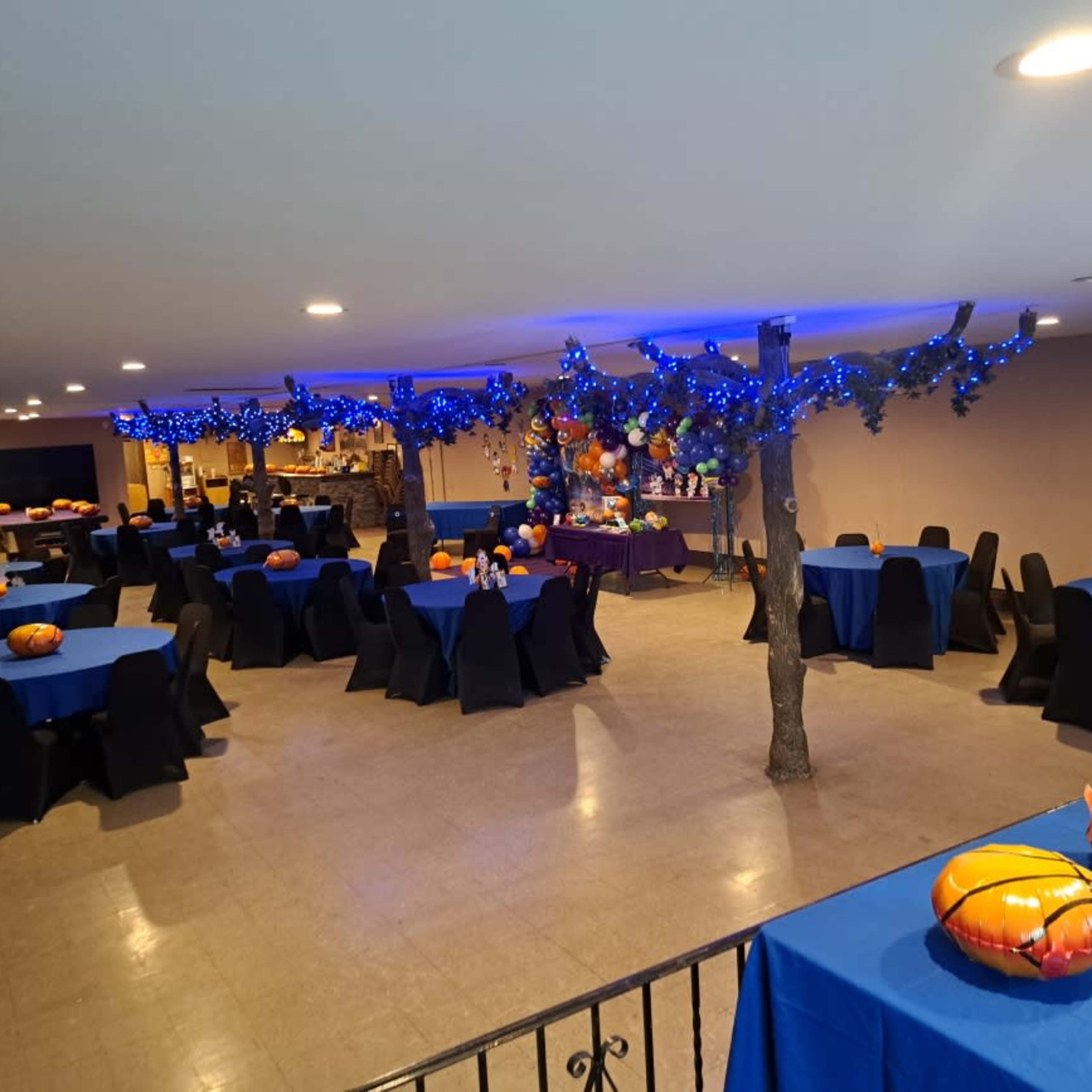 A decorated event space with blue tablecloth-covered tables, black chairs, and orange pumpkin centerpieces, all under blue string lights.