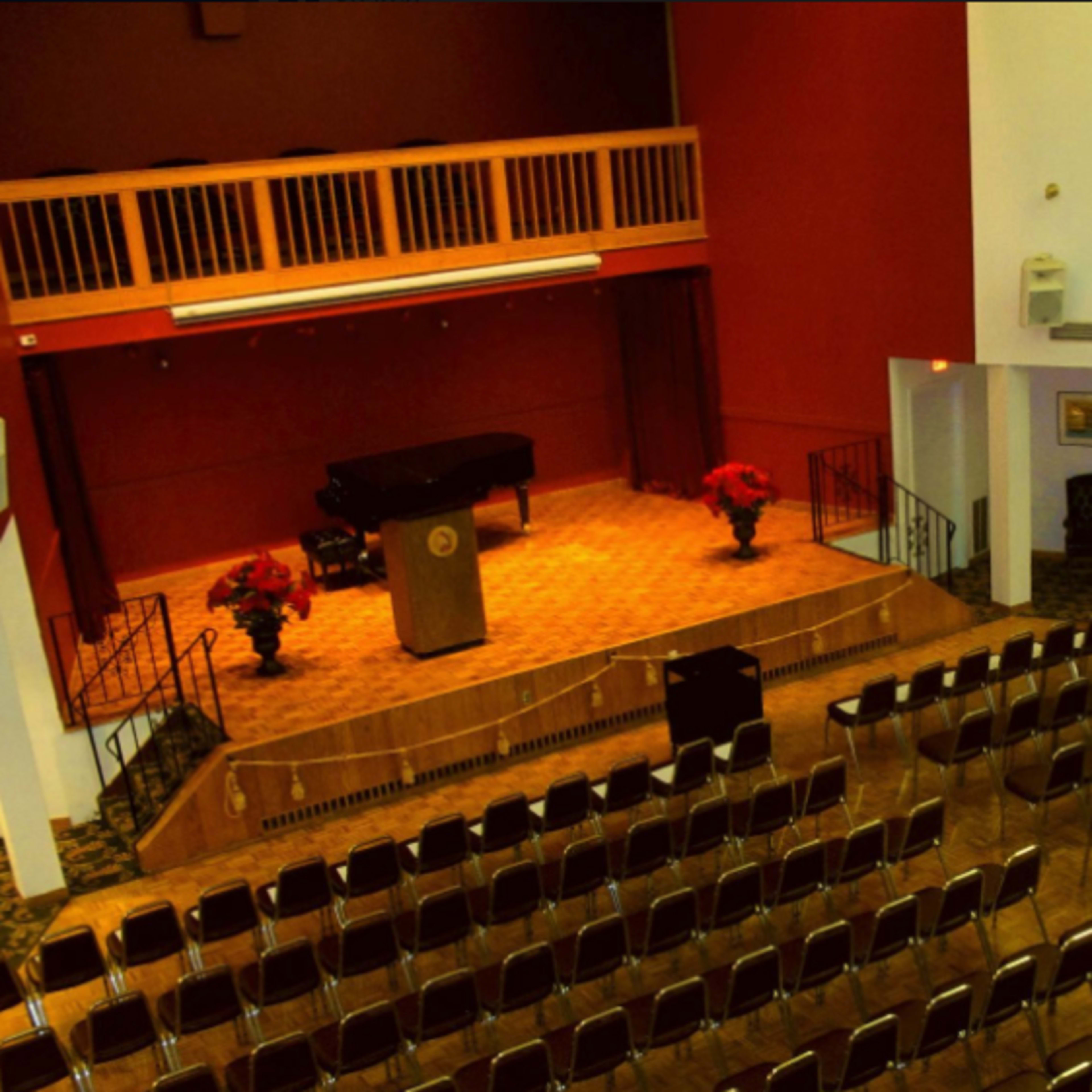 An auditorium with rows of empty chairs facing a stage that features a podium and a piano, flanked by potted plants.