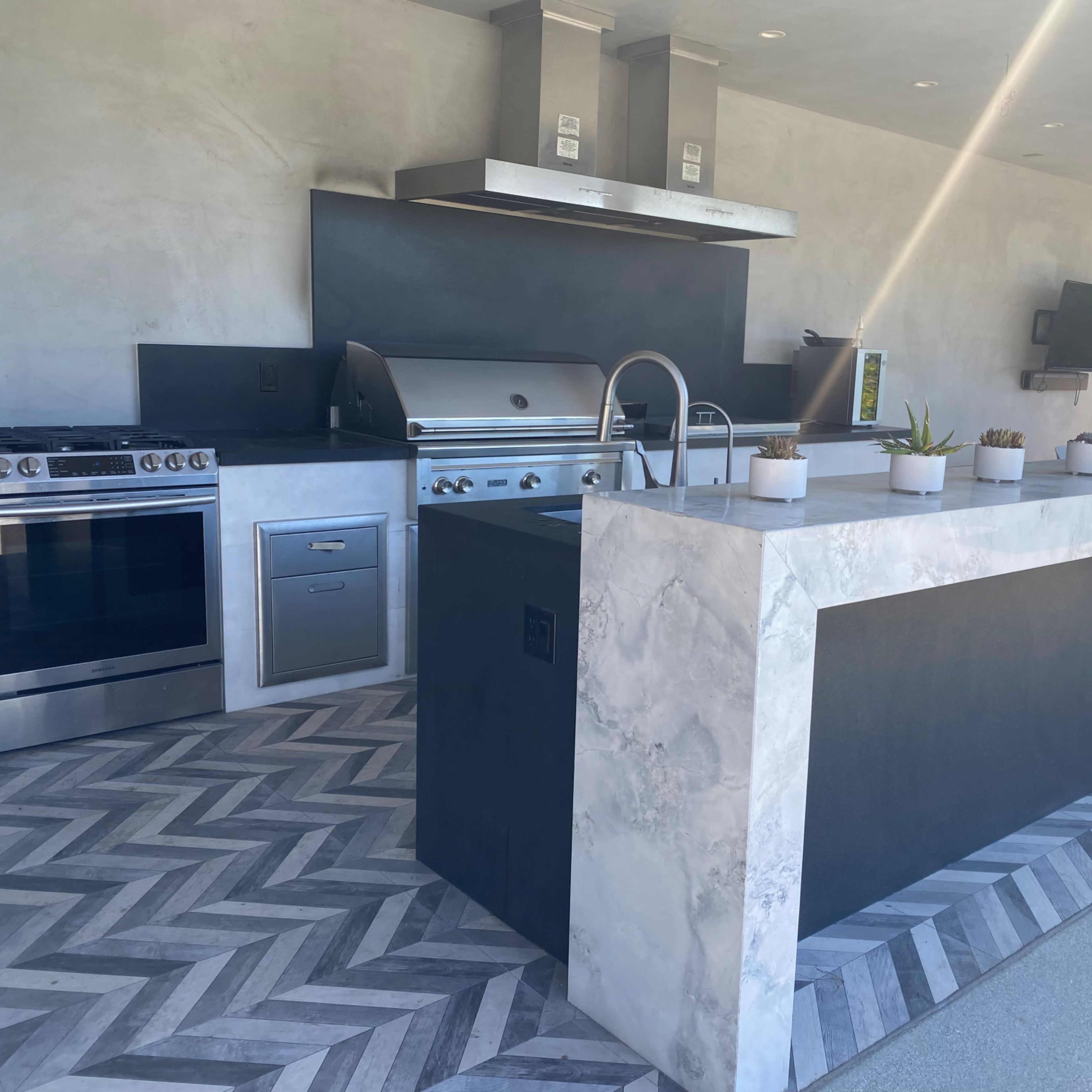 A modern outdoor kitchen featuring a large stone island, stainless steel appliances, and a chevron-patterned floor.