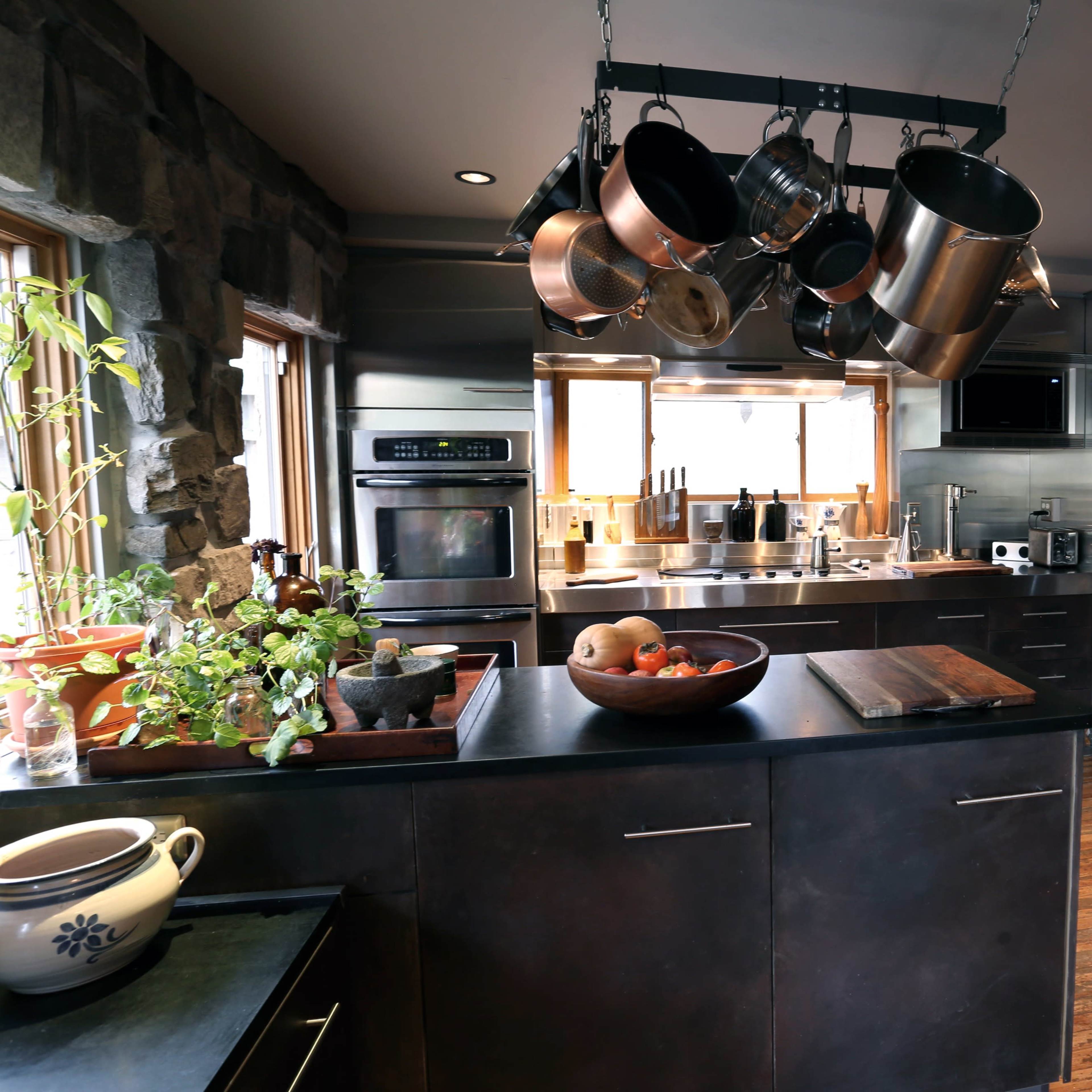 The kitchen features dark cabinetry, stainless steel appliances, and a hanging pot rack above an island with various kitchen utensils and a bowl of fruit.