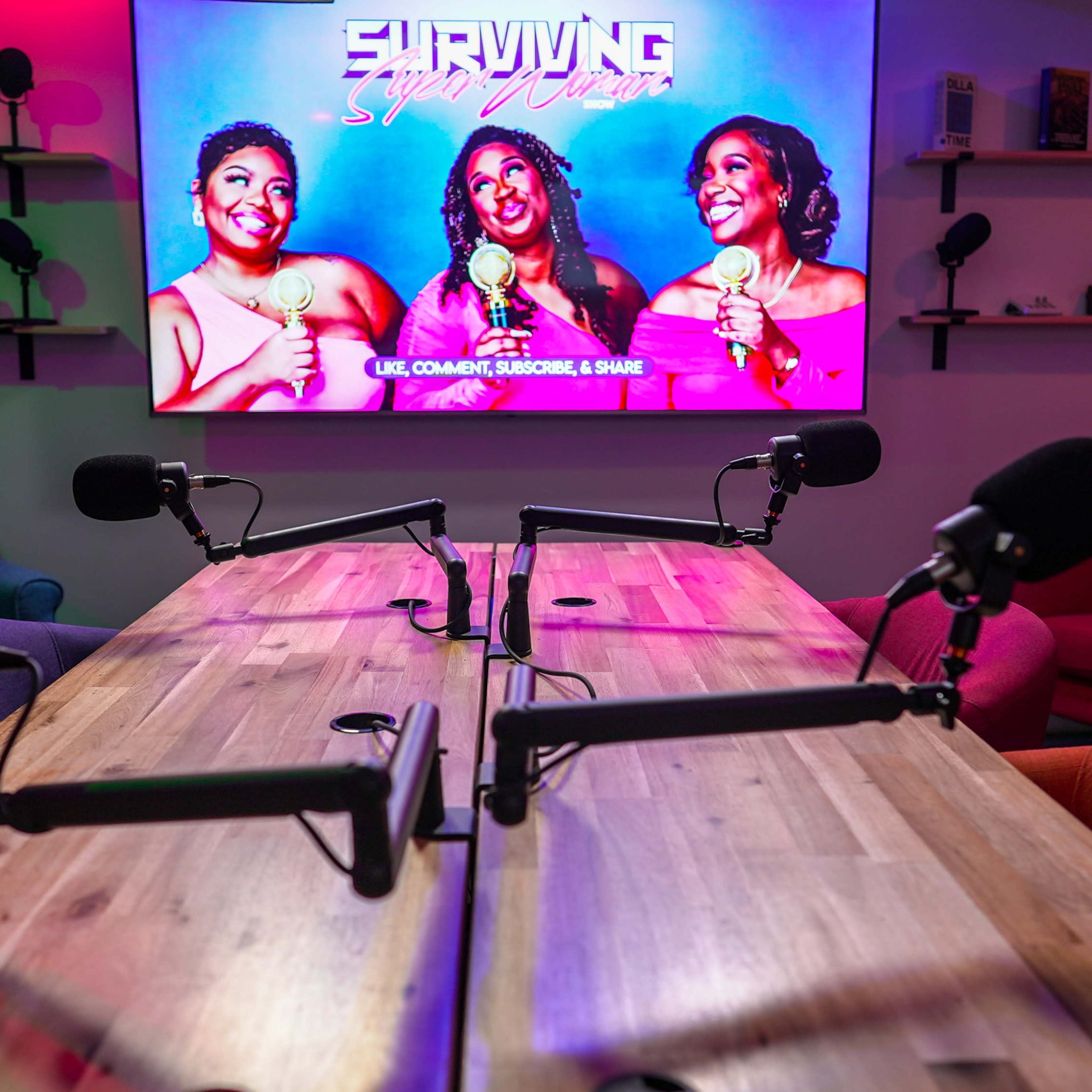 A recording studio setup with a wooden table, two microphones on each side, and a large screen displaying the title "Surviving Super Chicas."