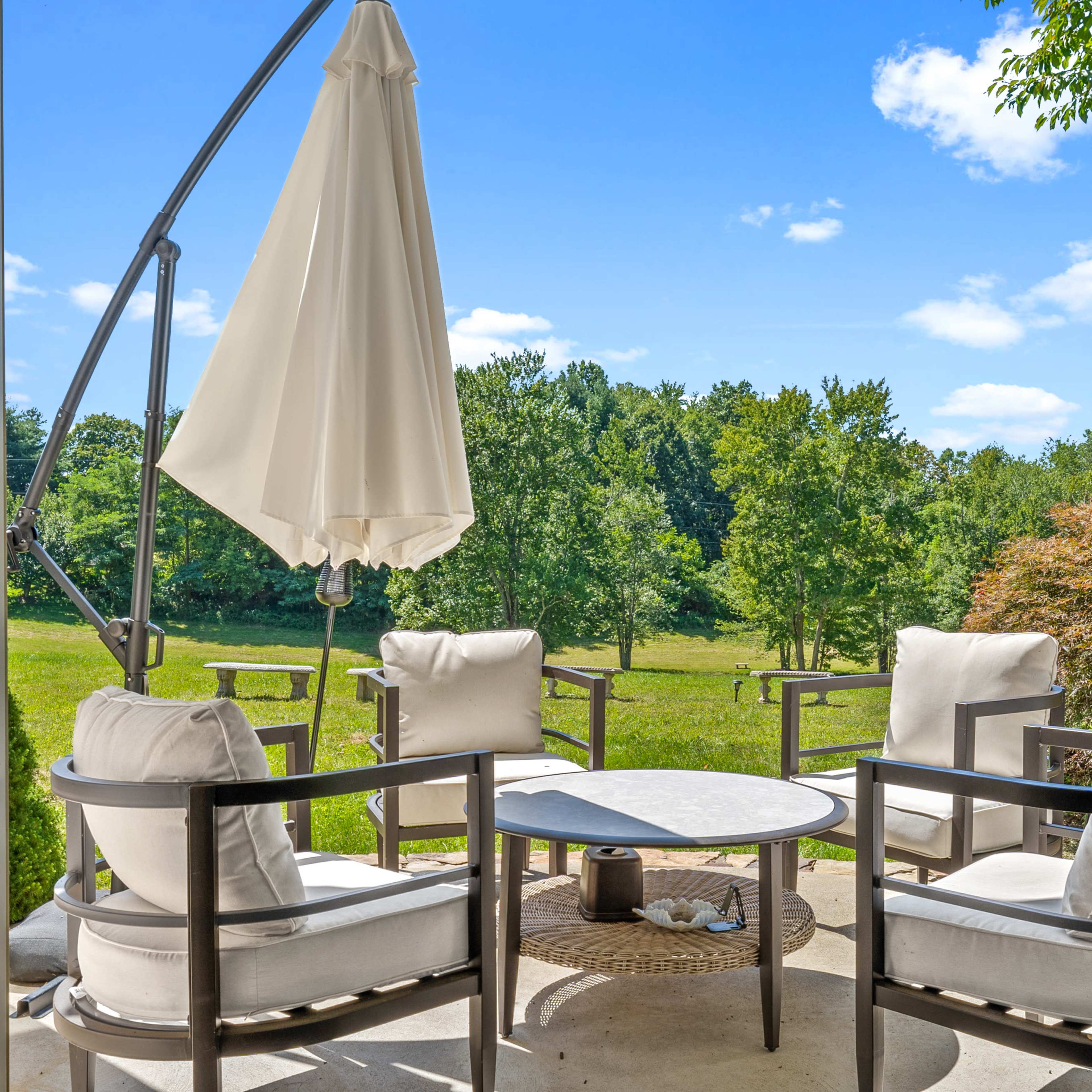 A patio scene features four cushioned chairs arranged around a small circular table, with a large umbrella poised above, overlooking a green landscape dotted with trees.