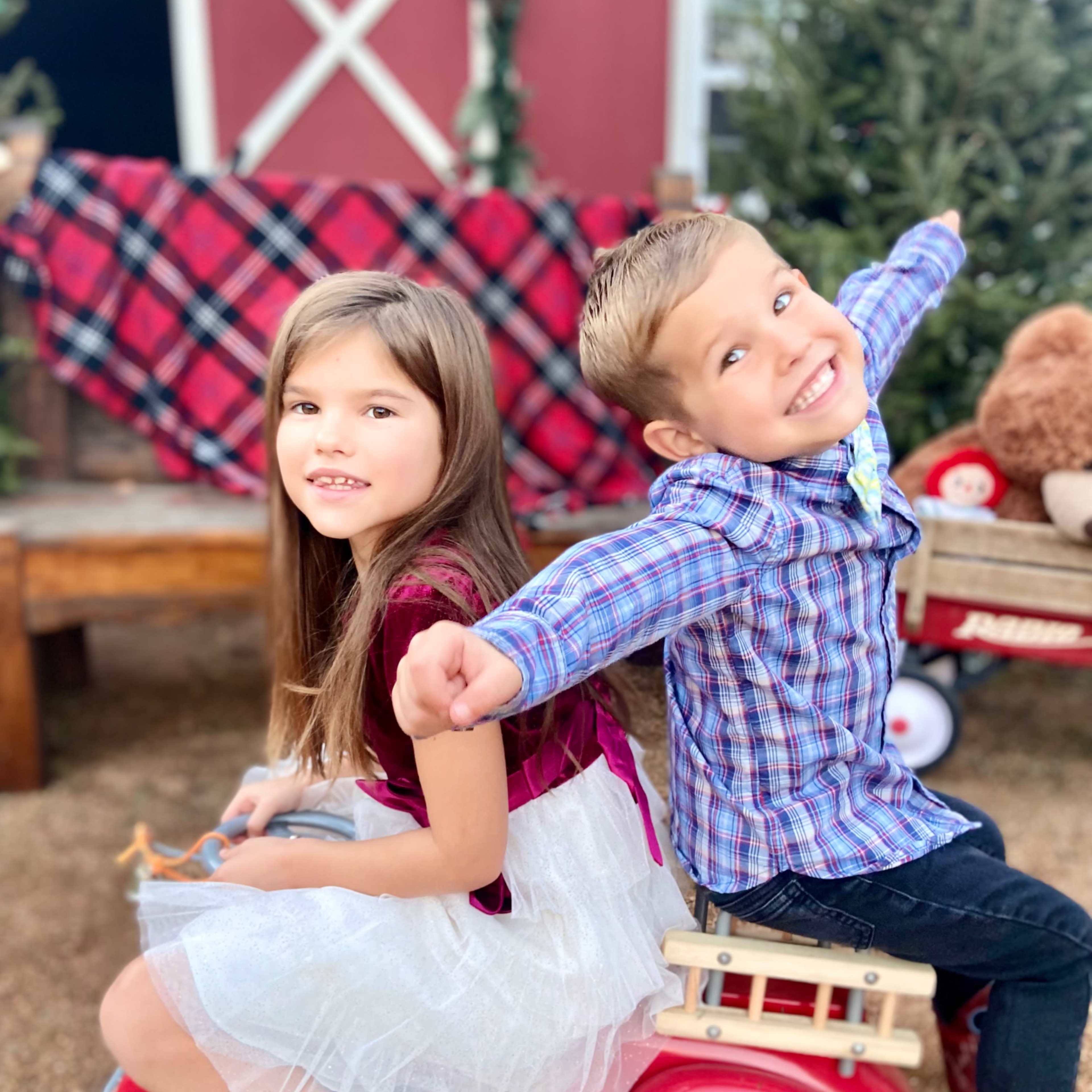 Two children sit back-to-back on a red toy car, with one girl wearing a white skirt and the boy in a plaid shirt smiling widely.