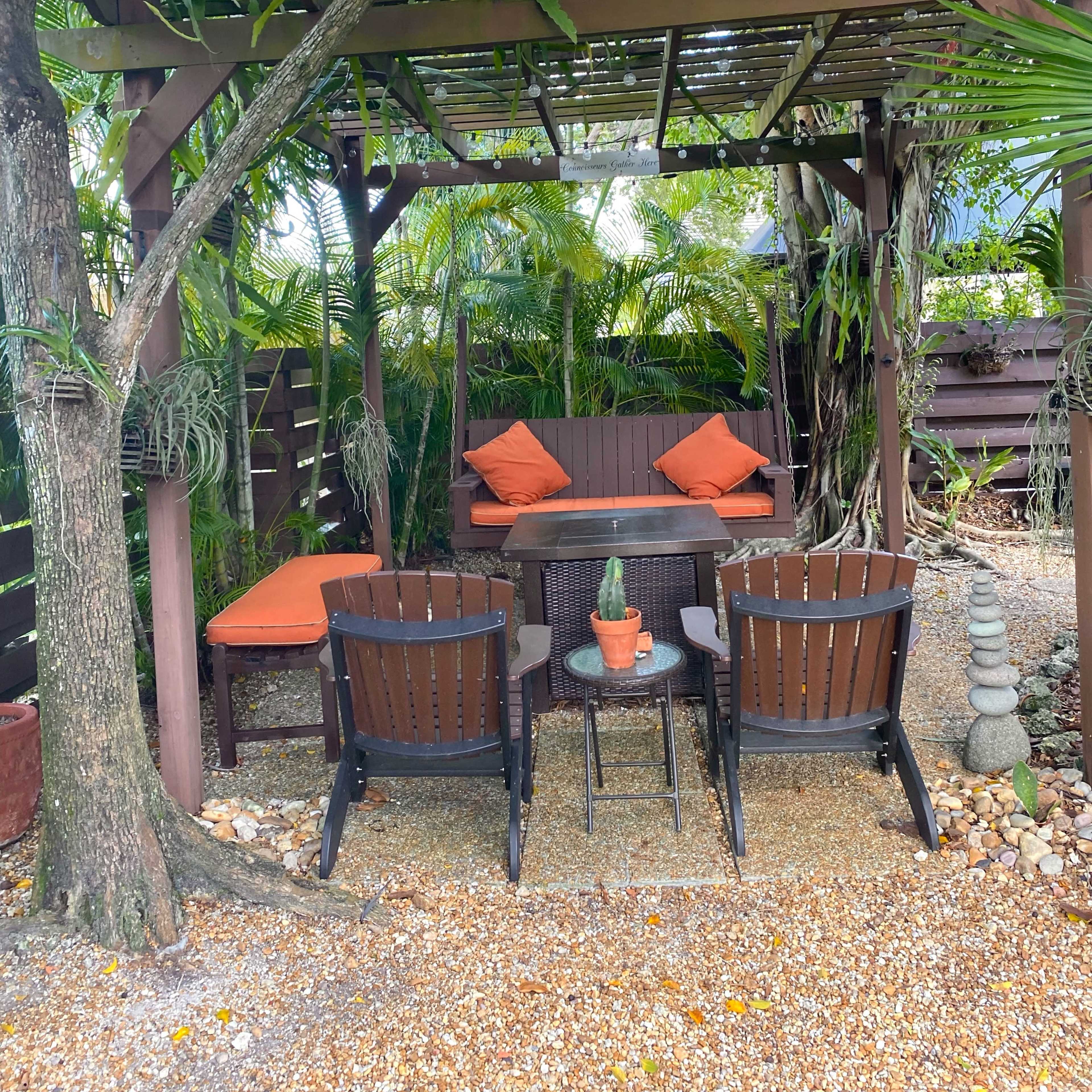 A shaded outdoor seating area features two wooden chairs, a small table with a potted plant, and cushions on a sofa surrounded by tropical greenery.