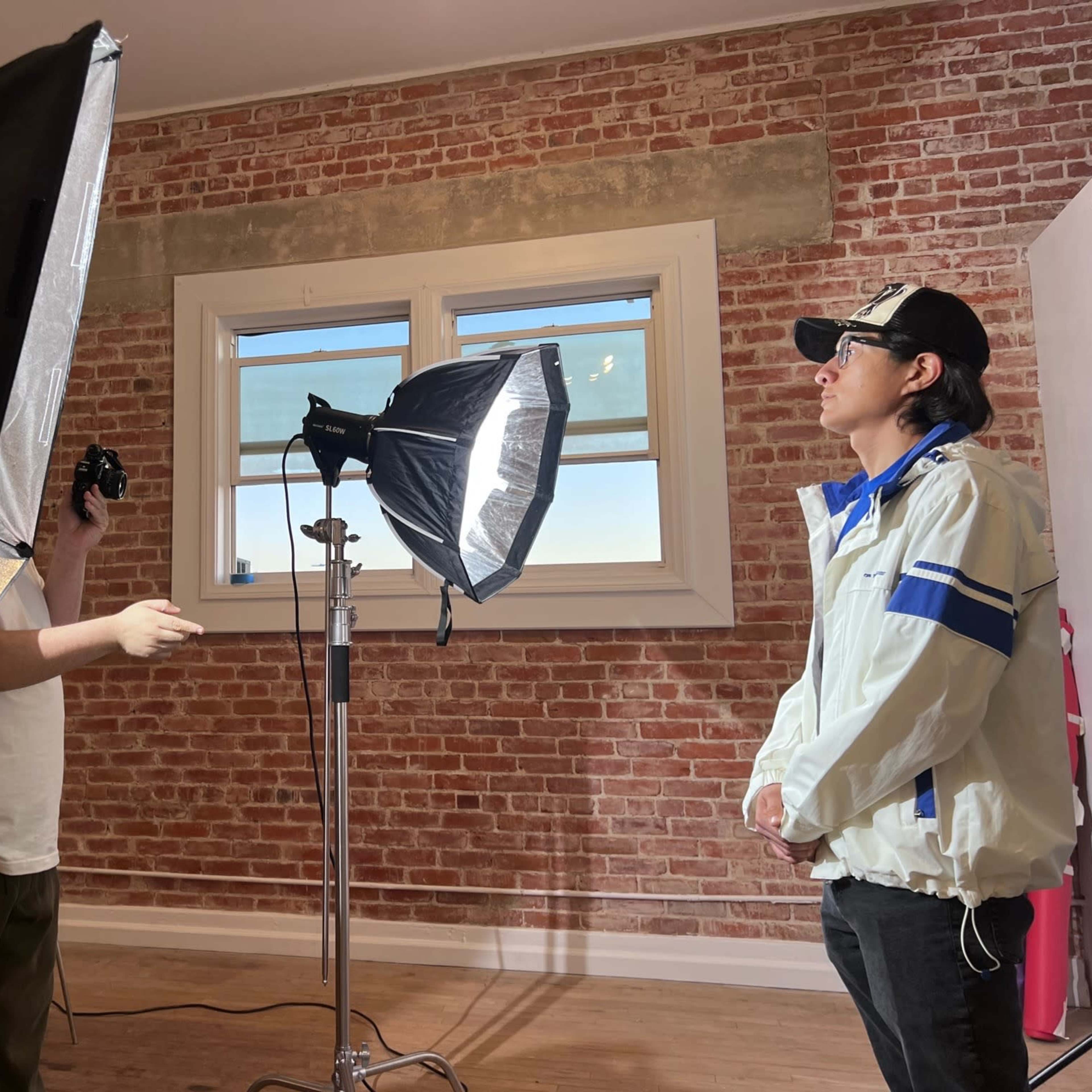 A person in a white jacket stands in front of two studio lights while another person operates a camera in a room with exposed brick walls.