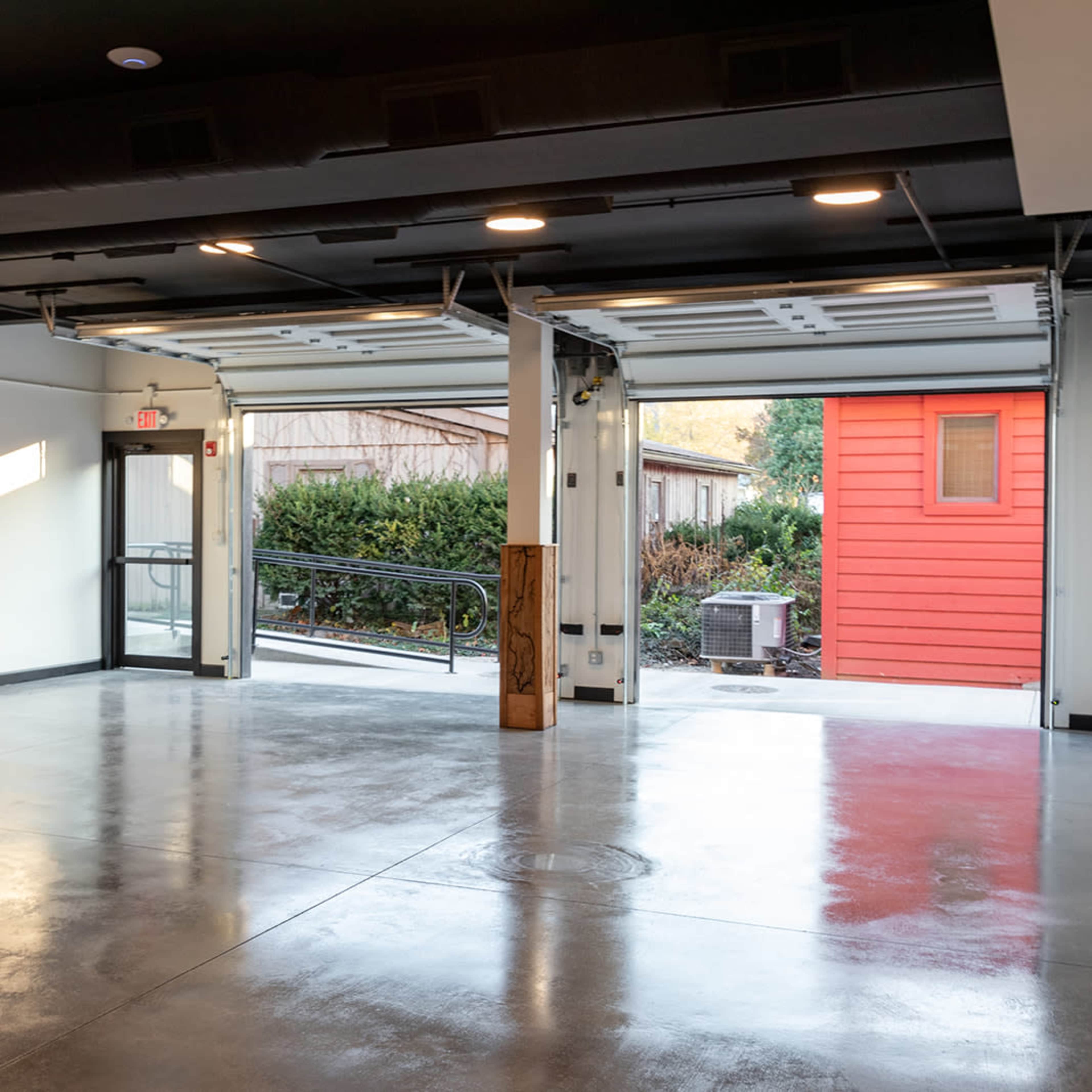 An empty industrial-style room with polished concrete floors and large, open doors leading to an outdoor area and a red structure.